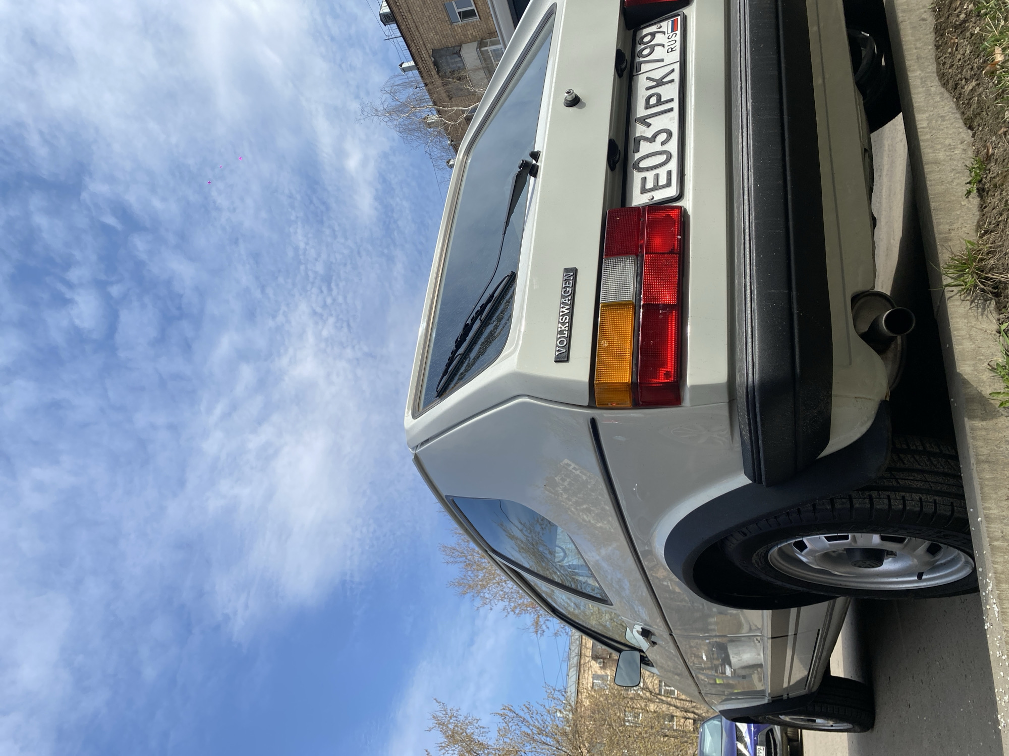 A white Volkswagen car is parked on a street in Khimki, Russia. The car's license plate reads "E031PK799 RUS". The sky above is blue with scattered white clouds. A building is visible in the background. The car is parked close to the curb.