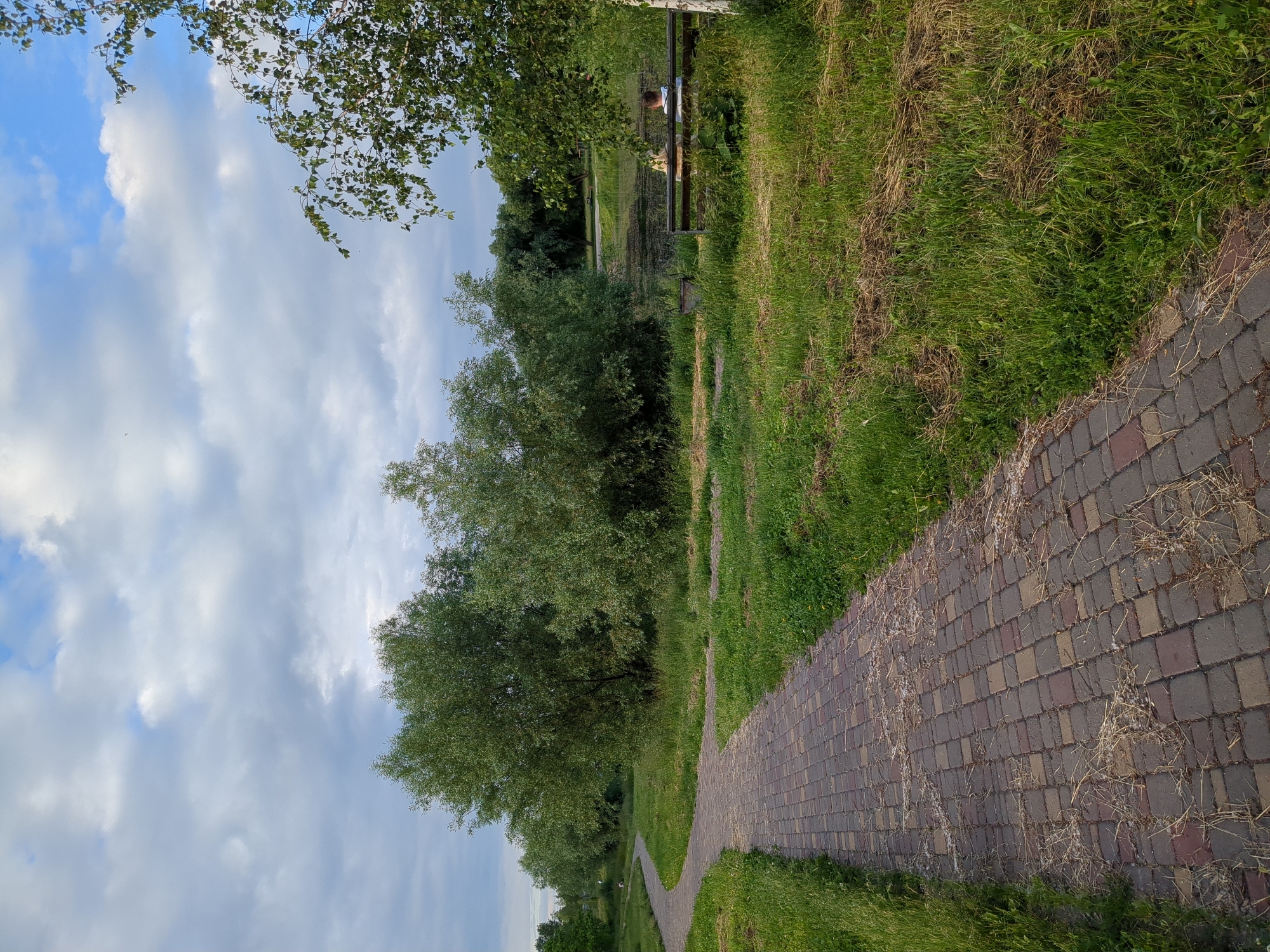 The image depicts a park scene in Boryspil, Ukraine. A paved pathway made of interlocking bricks curves through lush green grass. On the right side of the pathway, a wooden bench is occupied by two individuals. One person, with light-colored hair, is partially visible from the side, and another person wearing a white shirt is seated next to them. Beyond the bench, a body of water, likely a pond or lake, is visible, surrounded by dense green foliage and trees. The sky above is partly cloudy with patches of blue. The overall environment suggests a tranquil outdoor setting within the city.