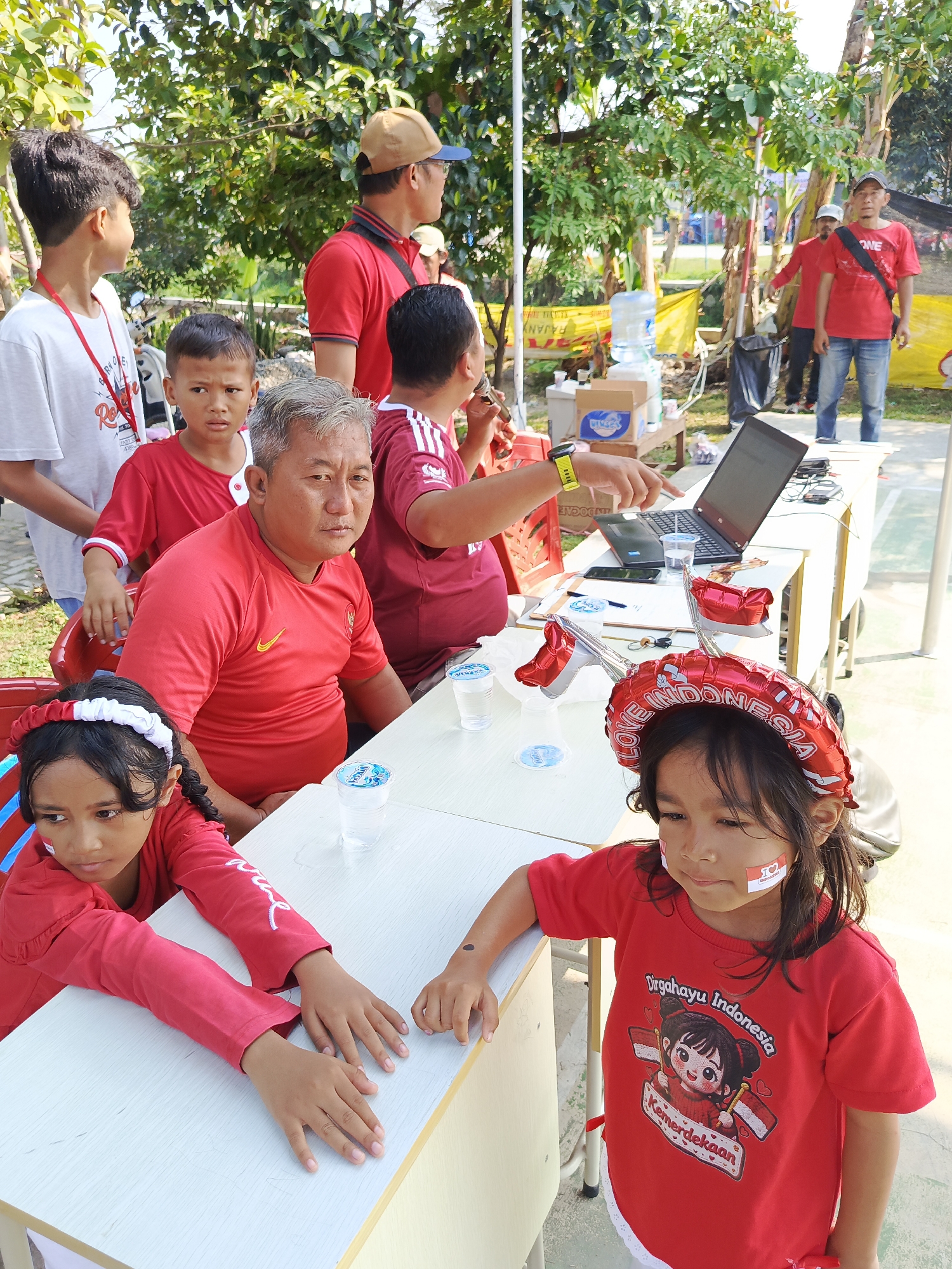 The image captures a daytime outdoor scene in Ciseeng, Indonesia. In the foreground, two young girls in red shirts are seated at a white table. The girl on the left wears a red and white headband and has what appears to be face paint on her cheeks. The girl on the right wears a red headband with "LOVE INDONESIA" printed on it, and also has face paint on her cheeks. Her red t-shirt displays a graphic with "Dirgahayu Indonesia" and "Kemerdekaan" text. Both girls are looking downwards. Behind them, several other individuals, mostly wearing red attire, are visible. An adult male with gray hair is seated at the table, looking towards the camera. Another adult male, holding a microphone or similar device, is standing behind him. In the background, more people, trees, and a yellow banner with Indonesian text are present. A table with a laptop and water bottles is also visible. The environment suggests an event or gathering taking place outdoors.