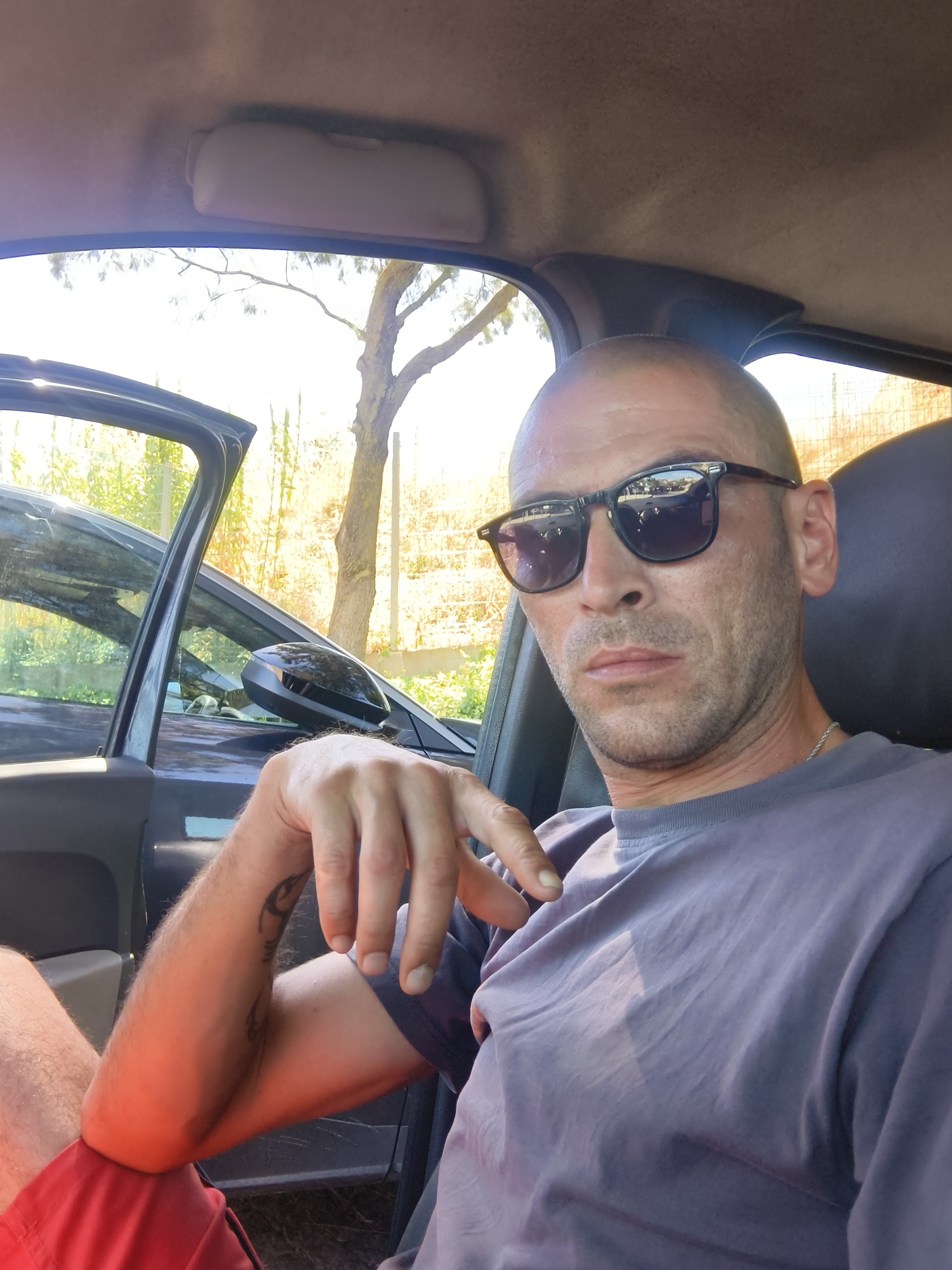 The image shows a man with a shaved head and sunglasses sitting in the driver's seat of a car. He is wearing a grey t-shirt and red shorts. His left arm is resting on his leg, and his hand is visible with a tattoo on his forearm. The car door is open, revealing trees and foliage outside. The scene appears to be outdoors, possibly in a rural or suburban setting, given the presence of vegetation and a fence in the background. There are no visible landmarks to pinpoint a specific location in Oeiras, Portugal.
