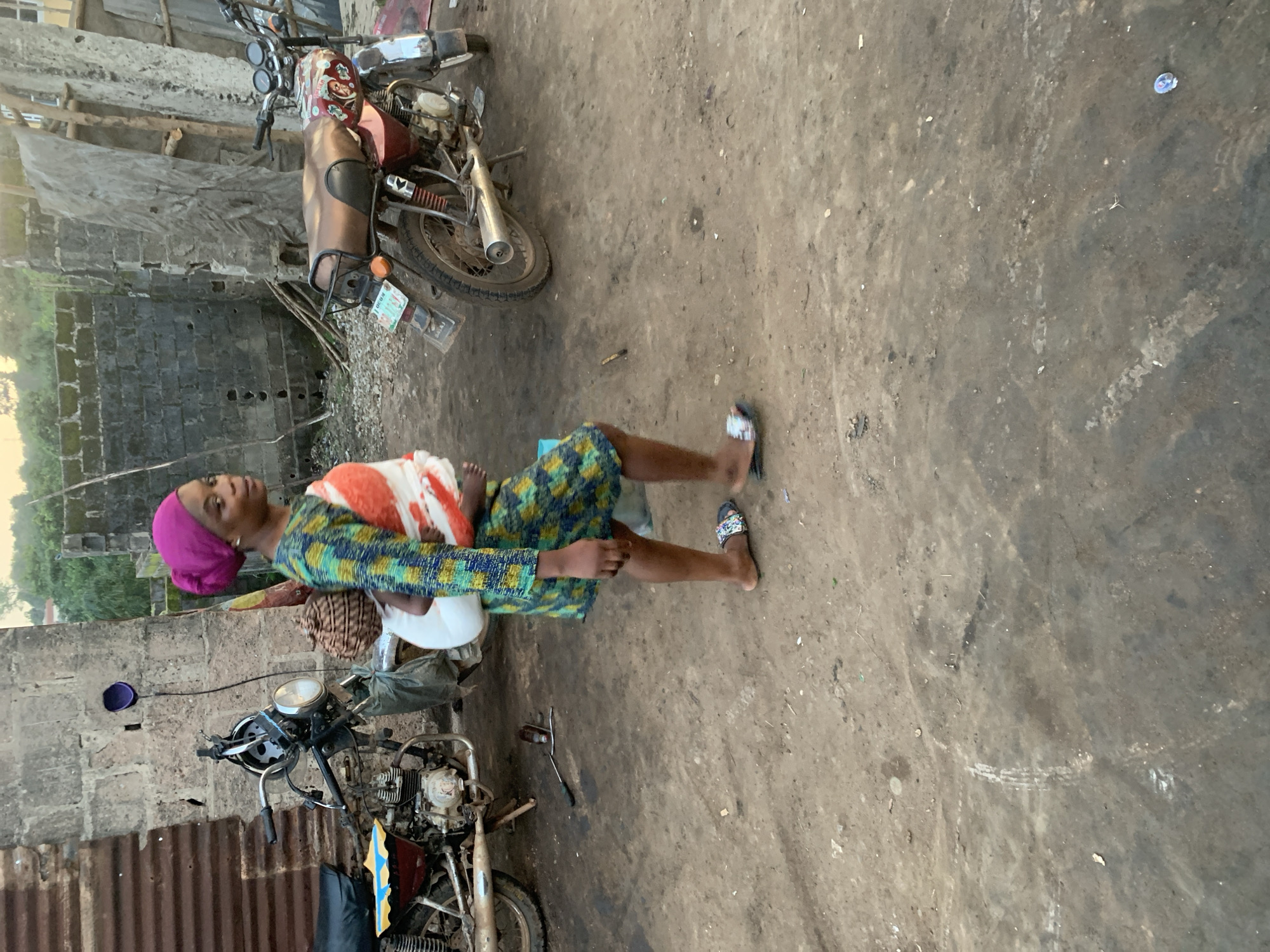 The image shows a woman wearing a vibrant, patterned dress and a pink headscarf, walking on a dirt ground in what appears to be an outdoor area of Shagamu, Nigeria. She is carrying a baby wrapped in a white cloth on her back, with a woven basket also strapped to her. To her left, a red and black motorcycle is parked next to a corrugated metal structure, and to her right, a burgundy motorcycle with a brown seat is visible. The background features a textured concrete wall and some wooden structures. The ground is uneven and appears to be a mixture of dirt and gravel.