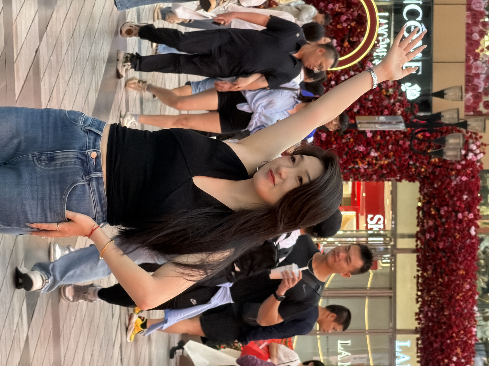 In Yuzhong District, China, a woman with long dark hair, wearing a black halter top and blue jeans, stands with her right arm raised, her hand open. She is positioned in the foreground, with her left hand in her jeans pocket. The background shows a blurred street scene with people walking. Decorative floral arrangements, predominantly red, hang overhead, and storefronts with neon signs are visible. The environment suggests a commercial or pedestrian area, possibly a shopping district.