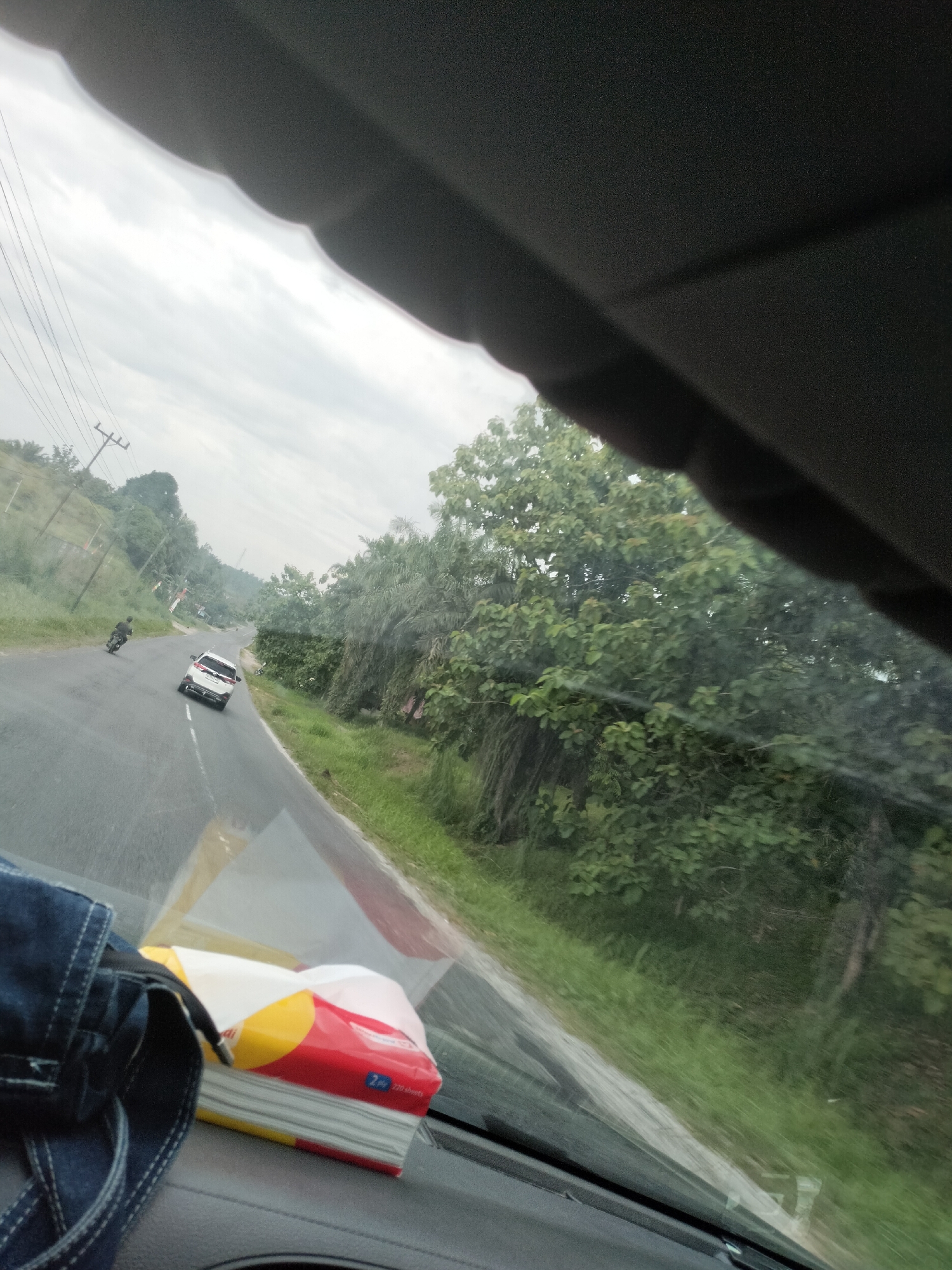 The image shows a view from inside a vehicle looking out at a rural road in Bukit Selamat, Indonesia. A white SUV is driving away from the camera on the asphalt road, which has a white dividing line. To the left of the road, there is a motorcycle with a rider, also moving away from the camera. The roadside is lined with green grass and trees. The sky is overcast. In the foreground, on the dashboard of the vehicle, a stack of facial tissues in a red and yellow package is visible, along with a pair of blue jeans. The overall impression is of a journey through a tropical or subtropical landscape.