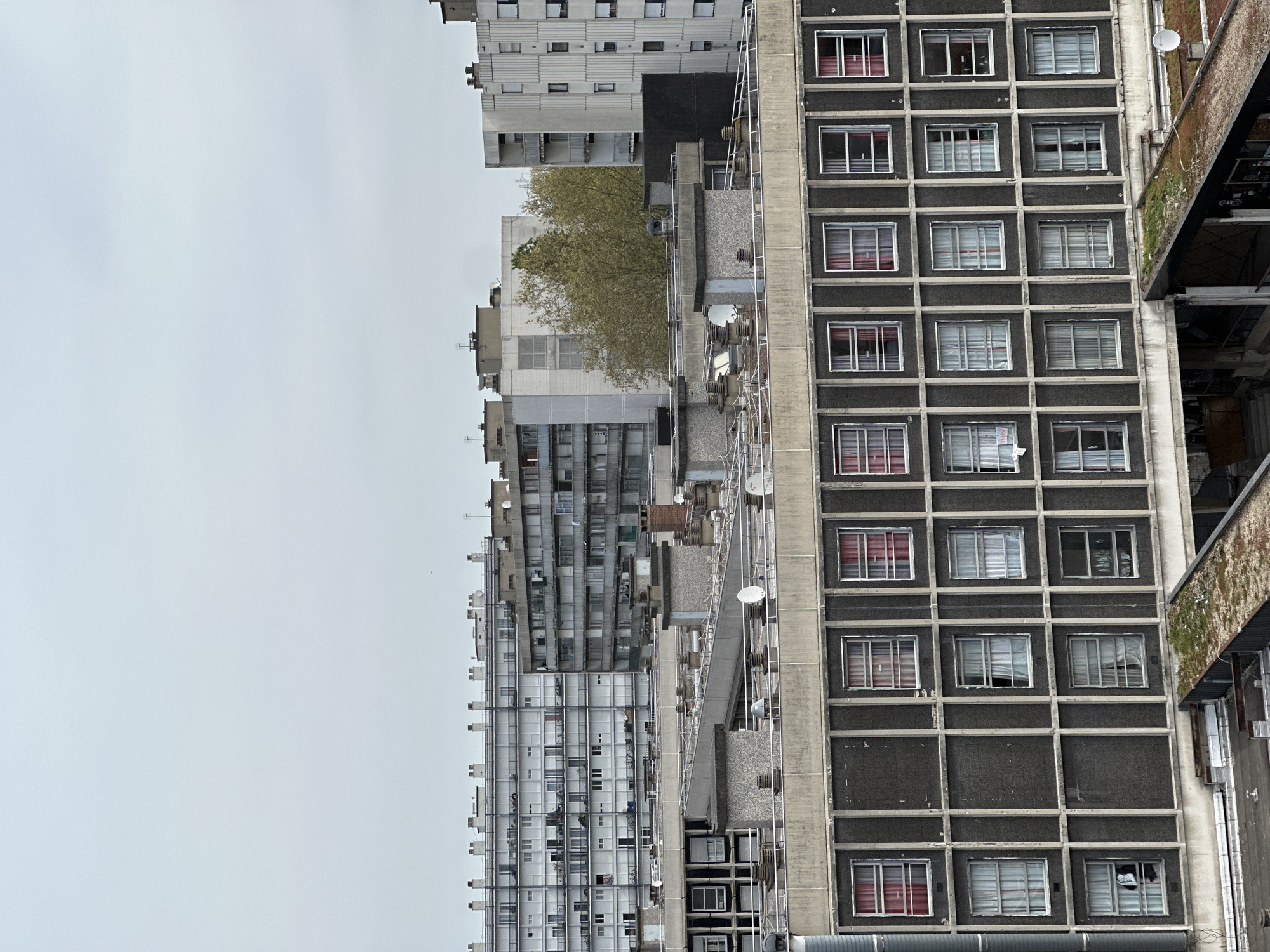 A medium shot presents a view of a group of high-rise apartment buildings on a cloudy day. The buildings are primarily gray, with some windows open and others closed. Satellite dishes are visible on the roofs of some buildings. The facade design of the buildings has a rough, repetitive pattern. A tree is placed in the middle. The scene appears to be located in Sarcelles, France, due to the architectural style and urban planning typical of French suburban areas. No people are visible in the frame. The overall impression is one of dense urban housing.