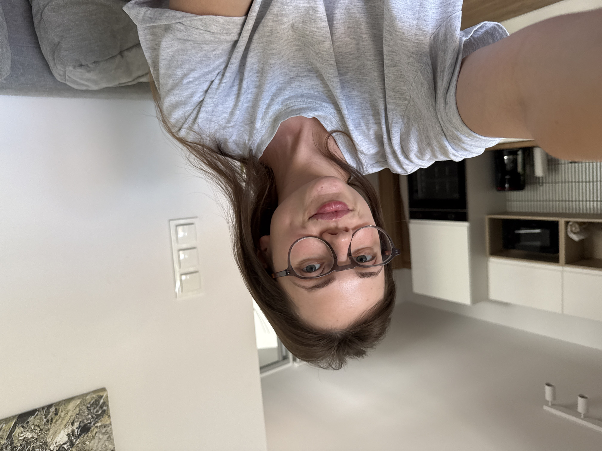 A woman with shoulder-length brown hair and glasses is captured in a selfie, facing the camera. She is wearing a grey t-shirt and appears to be indoors, possibly in an apartment. The background suggests a modern living space with white cabinets and kitchen appliances visible on the left side. A grey sofa is partially visible on the right. The lighting is natural and appears to be daylight. The woman's expression is neutral, with her lips slightly pursed. There is no indication of the specific location being Szklarska Poręba, Poland.