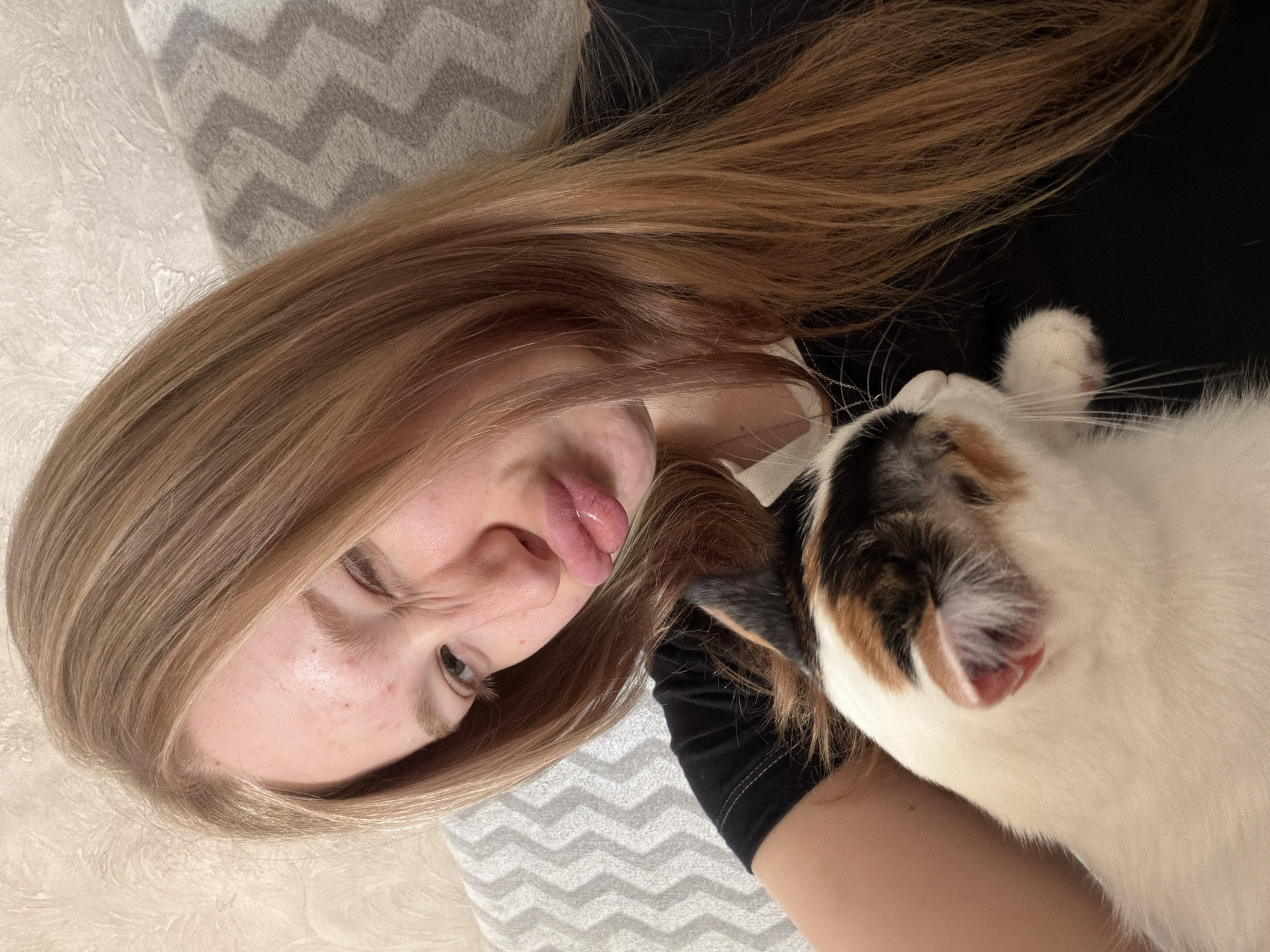 A young fair-skinned woman with long, light-brown hair is lying down with a calico cat. The woman is sticking her tongue out and looking to her left. The cat is mostly white with patches of orange and black, and is facing the right side of the frame. Both are lying on a patterned gray and white surface, possibly a bed. The woman is wearing a black top. This appears to be an indoor shot, with a second patterned pillow visible behind the woman's head. There are no specific details to identify this photo as being taken in Rossiyskiy, Russia.
