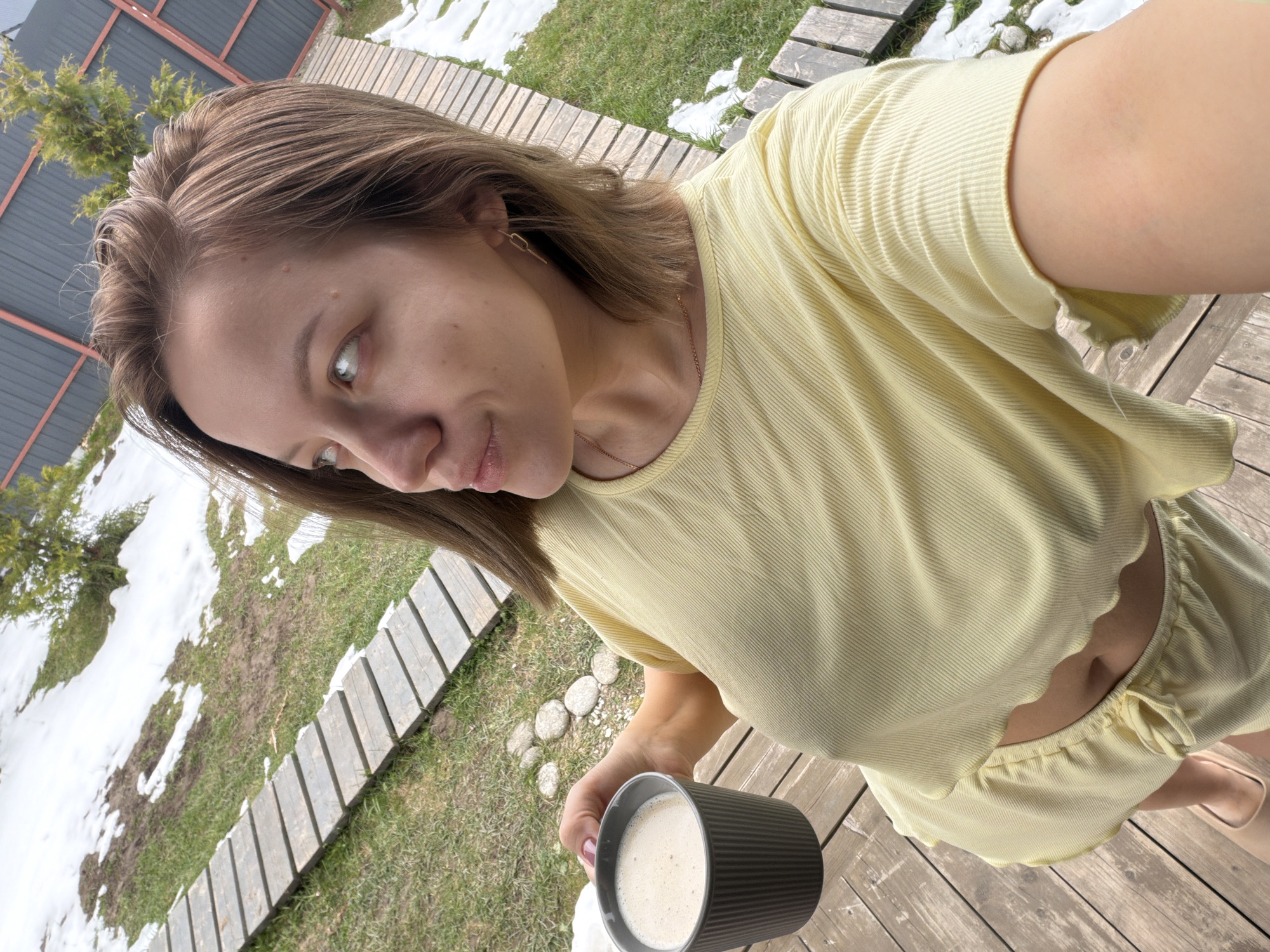 A young woman is depicted in a high-angle selfie, standing outdoors on a wooden deck in Бунятино, Russia. She has light brown hair and a fair complexion, and she is looking slightly away from the camera with a relaxed expression. She is wearing a light yellow, ribbed, short-sleeved top with a ruffled hem that exposes her midriff, and matching shorts with a drawstring waist. A delicate gold-colored necklace and a small hoop earring are visible. In her right hand, she holds a dark gray, ribbed mug containing a light-colored beverage, likely coffee or tea with milk.
The background shows an outdoor setting with a mix of green grass and patches of melting snow, suggesting early spring or late winter conditions. A wooden plank pathway extends across the grassy area. In the distance, a dark gray fence and some green conifers are visible. The scene is brightly lit by natural sunlight, indicating it is daytime. The overall impression is one of a casual, relaxed moment enjoying a drink outdoors. There is no visible text in the image.
