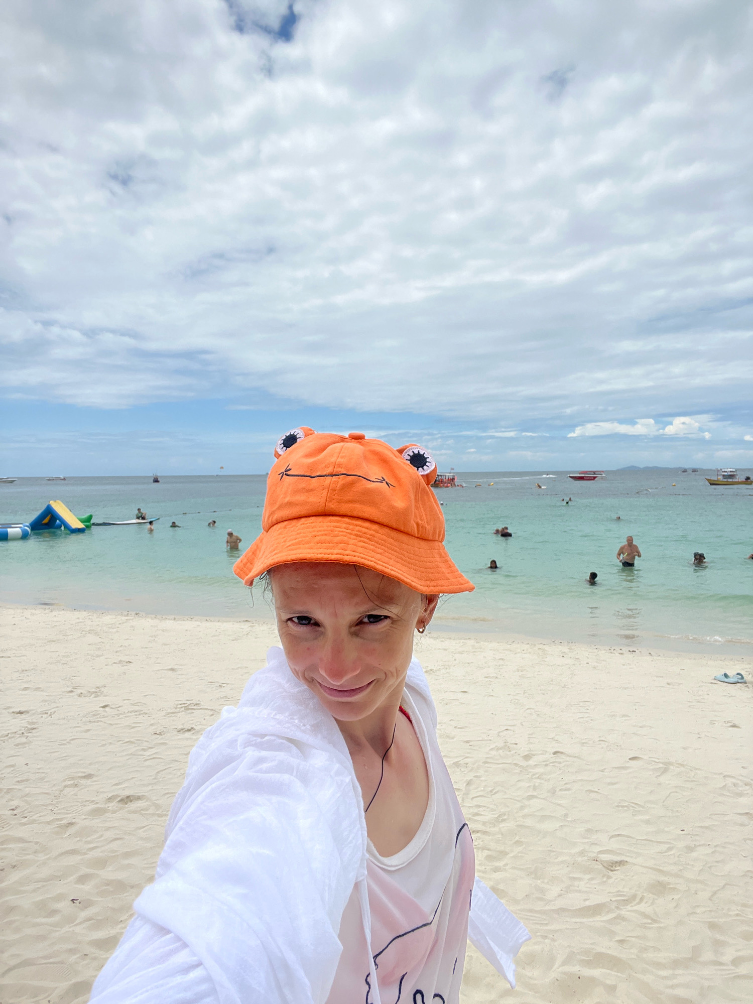 A medium shot shows a person wearing an orange hat with frog eyes, a white cover-up, and a white shirt with pink and black designs. They are on a sandy beach with the ocean in the background, located in Pattaya City, Thailand. The ocean is a light turquoise color and has several people swimming and floating on inflatable toys. Several boats are visible in the distance, as well as a parasailer in the sky. The sky is partly cloudy with scattered white clouds against a blue background.