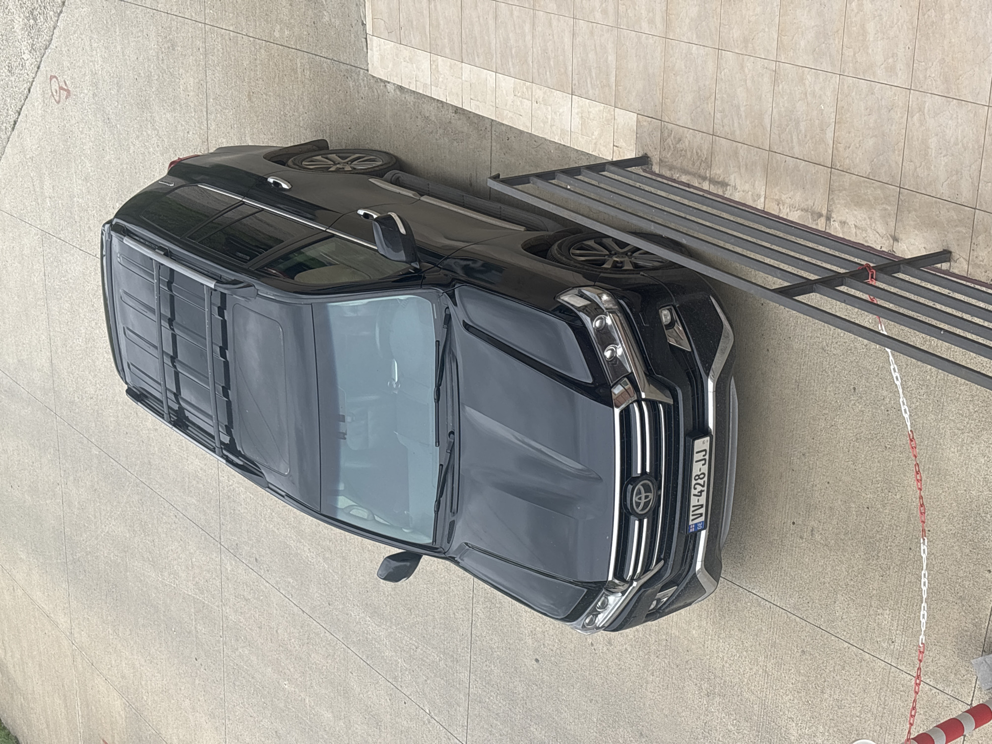 A black Toyota SUV is parked on a concrete surface near a building with tiled walls. A metal structure is situated next to the car. The SUV has a license plate reading "VV-428-JJ," which is a Georgian license plate. The presence of the Georgian license plate suggests the photo was taken in Tbilisi, Georgia. A red and white chain barrier is near the front of the vehicle. A disabled parking sign is visible on the concrete surface.