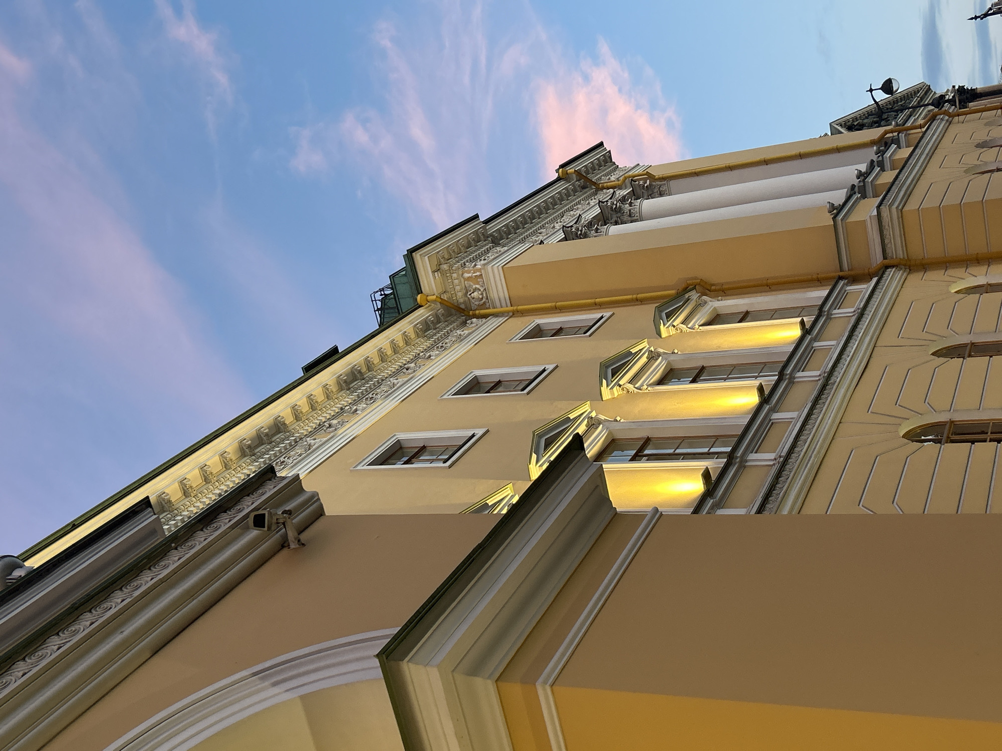 The image shows the exterior of a large, ornate yellow building with white architectural details. The building has multiple windows, some of which are illuminated by warm, yellow lights from above. The sky is a mix of blue and pink, suggesting it is either sunrise or sunset. The building's facade features classical architectural elements like columns and detailed friezes. The perspective is from a low angle, looking up at the building. A security camera is visible mounted on the building's exterior near the roofline. A lamppost with a single globe light is also visible on the right side of the building. The overall impression is one of grandeur and historical architecture.