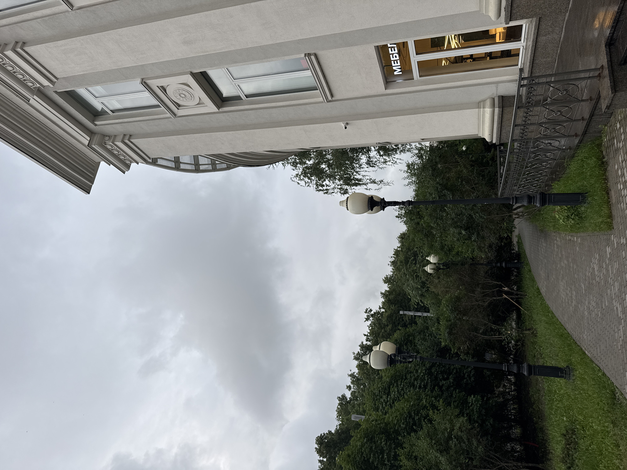 The image shows a light-colored building with classical architectural elements, including columns and decorative molding, on the right side. A sign with the Cyrillic word "МЕБЕЛ" (MEBEL, meaning furniture) is visible above a storefront window. To the left of the building, a paved walkway curves through a green, tree-lined area with lampposts. The sky is overcast with gray clouds. The scene appears to be an urban environment, possibly a street or park area within Moscow, Russia.