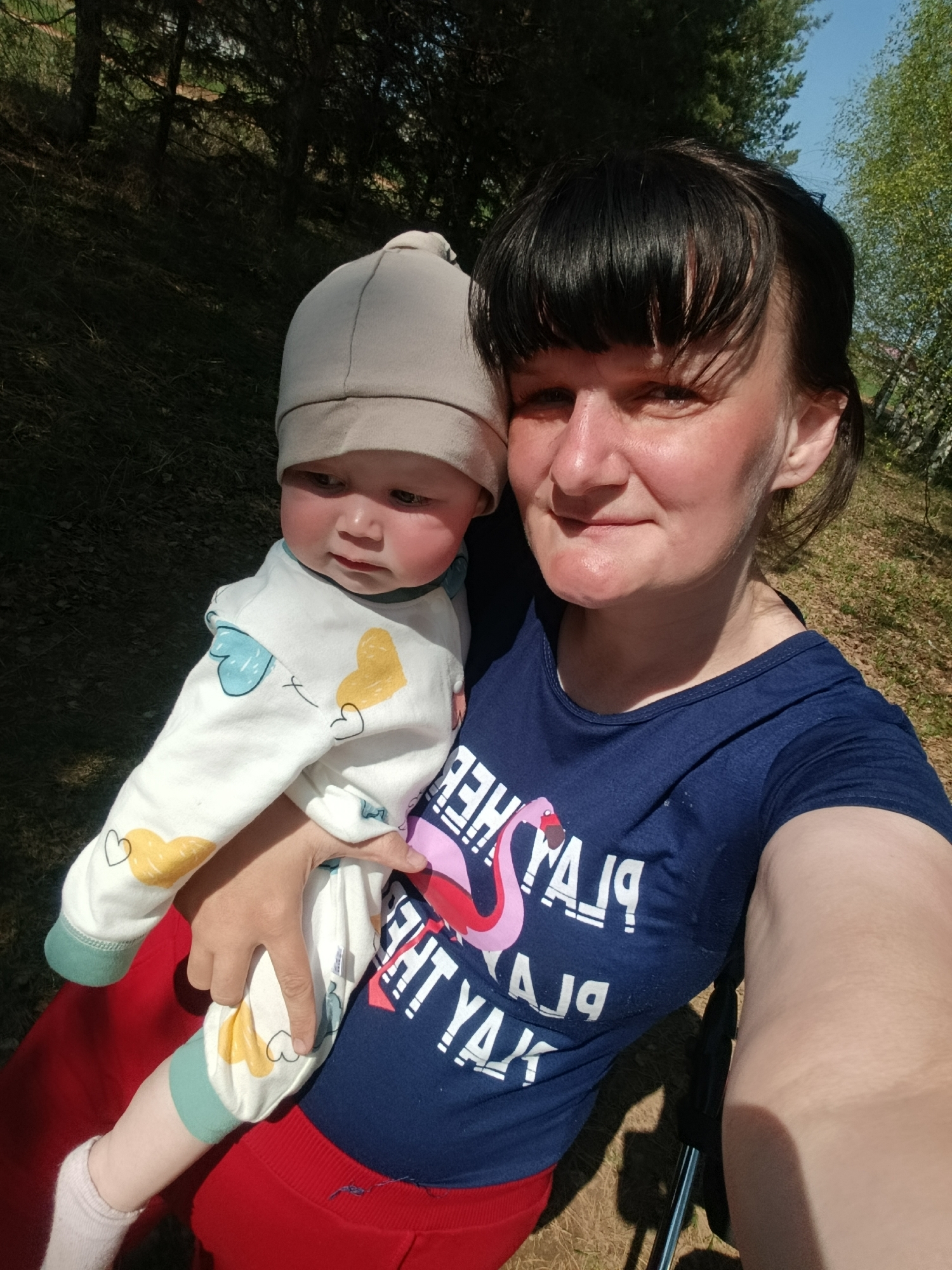 A woman in a navy blue t-shirt with a flamingo design and the words "HERE TO PLAY, PLAY YA JAY" is holding a baby wearing a light beige hat and white onesie with colored hearts. The baby is looking to the left. The woman is smiling slightly at the camera, holding the baby with her left arm. The background is a blurry mix of green trees and grassy areas, suggesting a park or outdoor location. The woman is wearing red pants. The woman's right arm is visible and extended as if taking a selfie. The location cannot be verified as Фоки, Russia from the image.