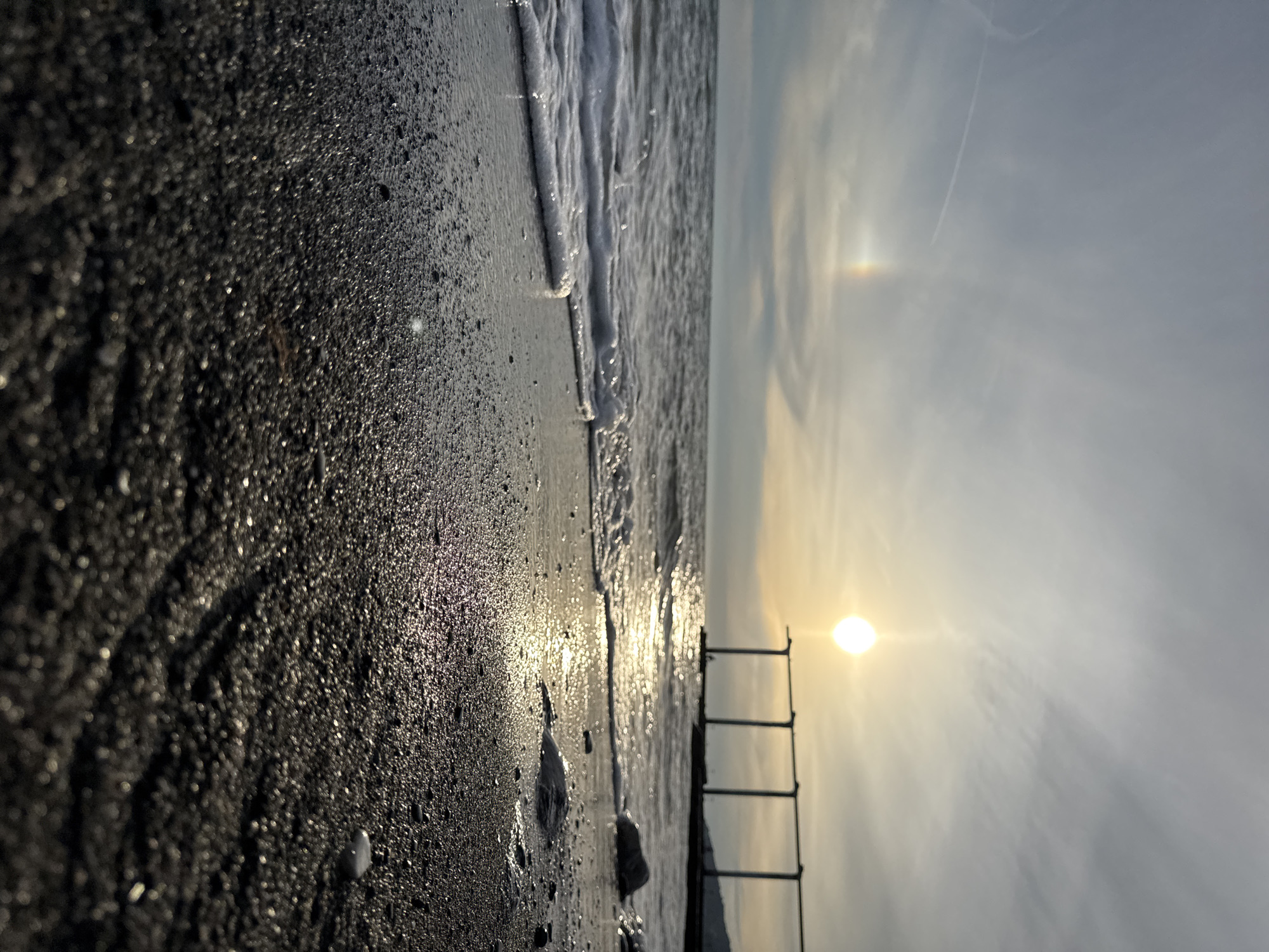 The image depicts a beach scene with the sun low in the sky, casting a warm glow on the water and wet sand. The sun appears as a bright orb with a halo, possibly indicating a sundog or atmospheric phenomenon. A partial rainbow is visible in the sky to the left of the sun. In the background, a weathered structure, resembling part of a pier or an old construction, stands in the water. The foreground is dominated by dark, wet sand with small pebbles and ripples. Gentle waves are washing onto the shore, creating a foamy edge. The overall mood is serene and tranquil, suggestive of a sunset or sunrise. The specific location in Sochi, Russia, cannot be determined from the visual information alone. No people are visible in the scene.