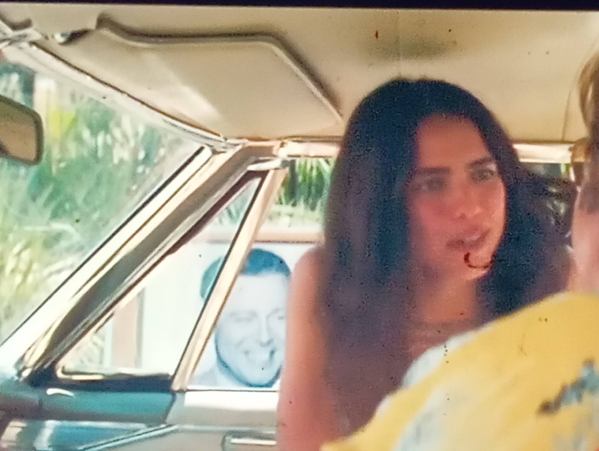 The image shows a close-up view from inside a car, looking out through the windshield and side window. The primary focus is on a young woman with long, dark hair, seen from the shoulders up, her face turned slightly away from the camera and her expression unreadable due to the blurriness. She appears to be in the passenger seat.
Through the side window, a man's face is visible, smiling broadly and looking out. He seems to be in the background or perhaps in another vehicle or building. The background outside the car is blurred, but appears to be foliage, suggesting an outdoor setting. The interior of the car is visible, including a portion of the roof lining and a sun visor. The overall lighting suggests it is daytime.
There is no visible text in the image. The image quality is grainy, suggesting it may be a still from a video or an older photograph. The context provided is Sofiivska Borshchahivka, Ukraine.