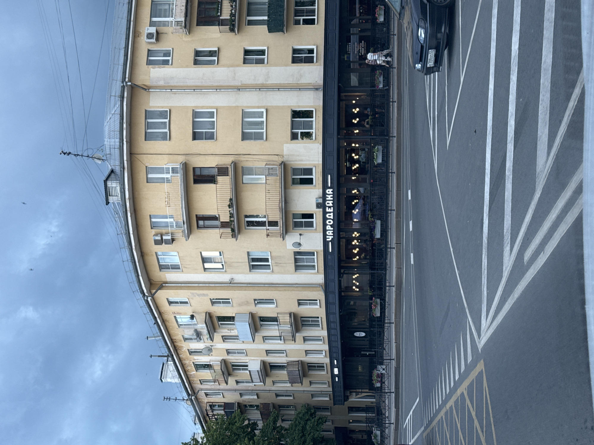The image shows a curved building in Veliky Novgorod, Russia, with a light yellow facade and white trim. The building has multiple stories with balconies on the upper floors. The ground floor features a dark-colored storefront with large windows, inside which lighting and people are visible. A sign above the storefront reads "ЧАРОДЕЙКА". The building is situated on a street corner with pedestrian walkways and road markings, including a crosswalk. A dark-colored car is visible on the right side of the image, driving past the intersection. The sky above is overcast with a cloudy, blue-gray appearance. Trees are visible to the left of the building.