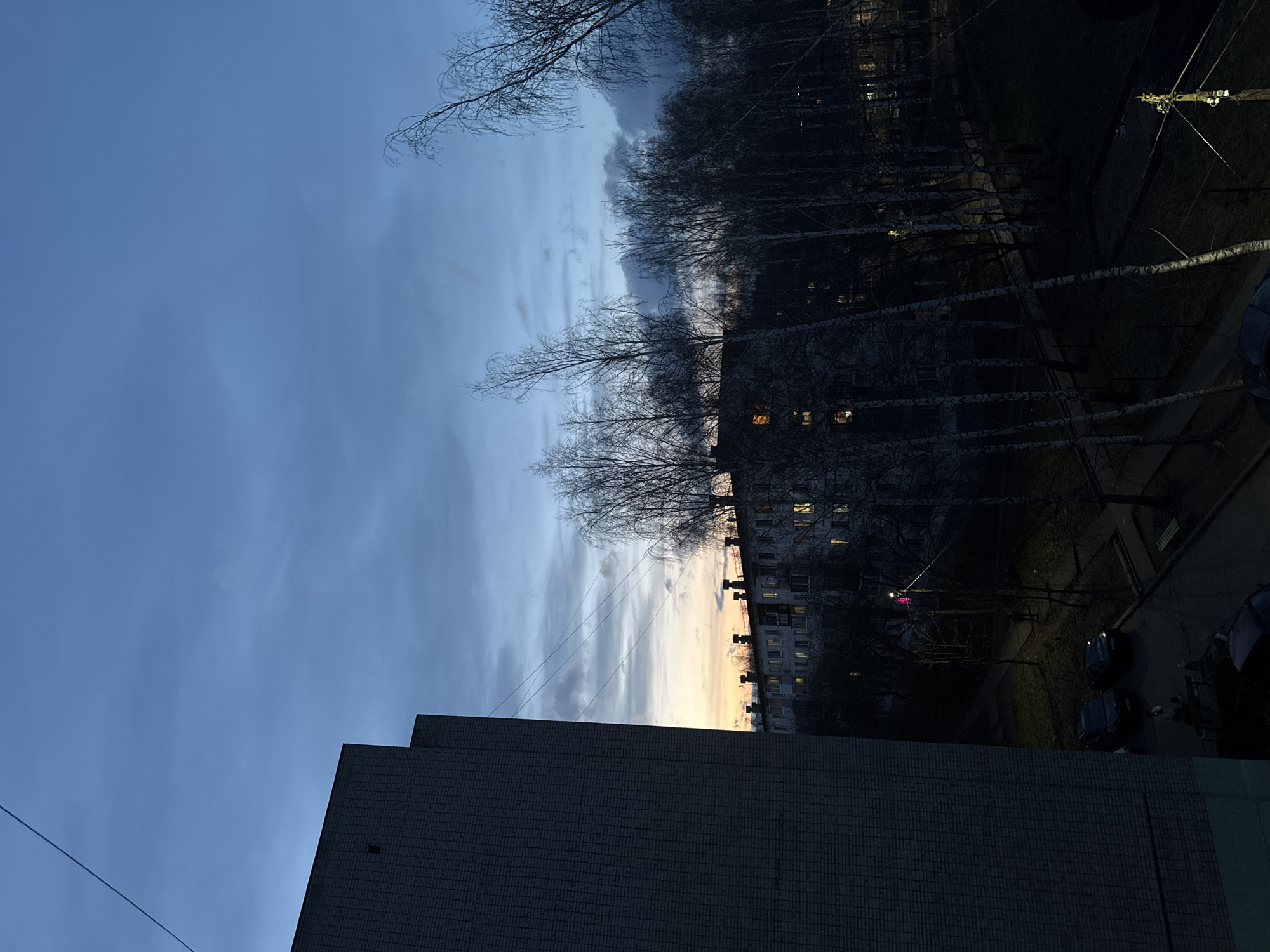 The image depicts an urban or suburban scene viewed from an elevated perspective, likely at dusk or dawn. The sky transitions from a deep, clear blue at the top to lighter tones, culminating in a warm orange and yellow glow near the horizon, indicative of either sunset or sunrise. Streaky clouds are visible across the sky.
Numerous bare, deciduous trees, possibly birches, form prominent silhouettes in the mid-ground, their intricate branches stark against the sky. Behind these trees, a multi-story residential building, characterized by illuminated windows, hints at human presence within. The architectural style appears consistent with buildings found in post-Soviet cities.
In the foreground, a dark, blocky building with a textured, tiled facade partially occupies the bottom-right of the frame. The ground level below the trees and buildings is dark, showing a paved area with two parked cars. Thin power lines traverse the upper sections of the image. A distant bright pink light source is faintly visible through the trees.
The scene conveys a quiet, contemplative atmosphere. The bare trees suggest the season is late autumn, winter, or early spring. No direct human activity is visible, and there is no discernible text in the image. The location context is Chaikovsky, Russia.