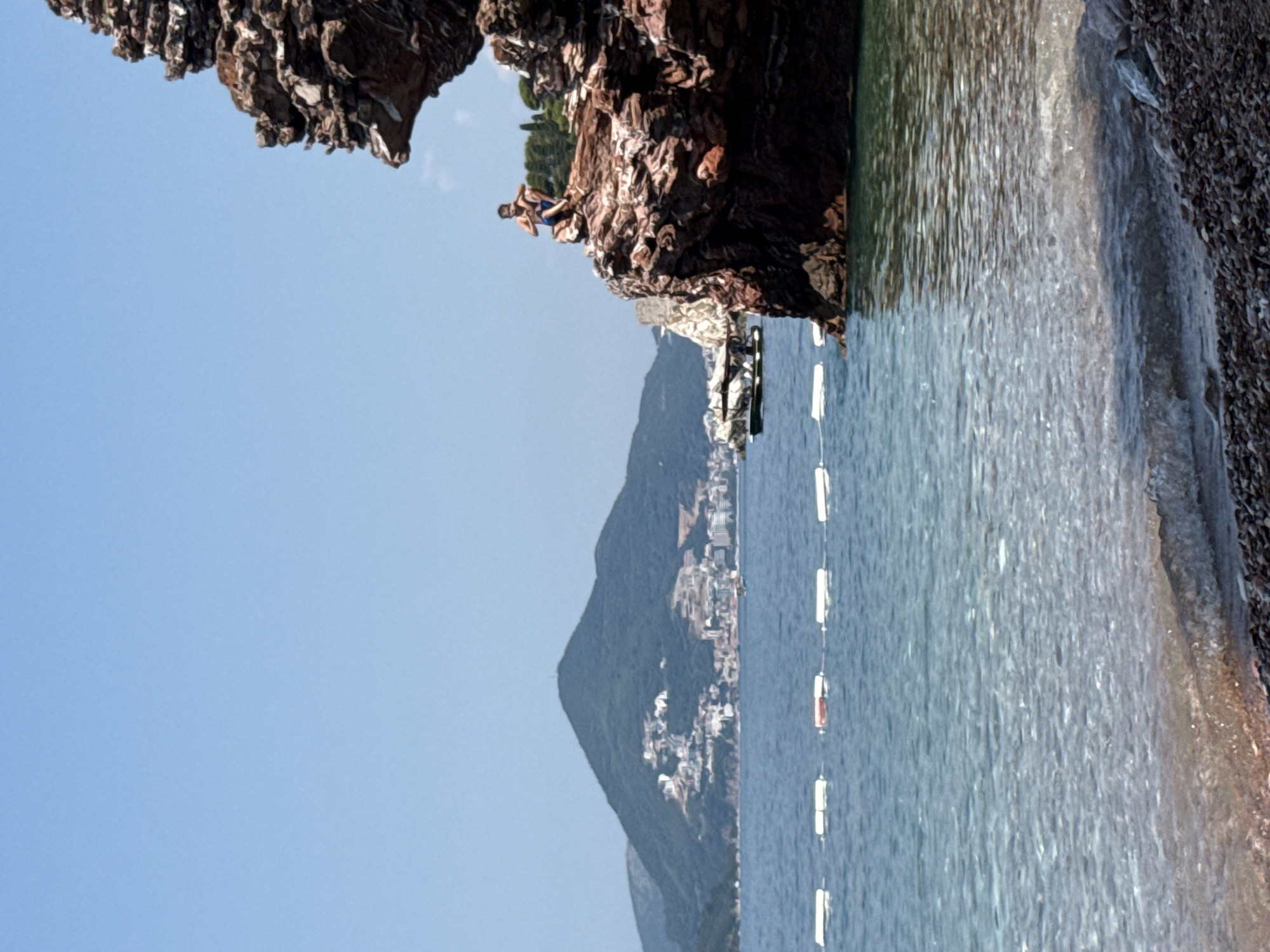 The image captures a dramatic coastal scene with a person perched precariously on a rocky cliff overlooking the Adriatic Sea. The perspective is from the water, looking up towards the cliffs. A young man, appearing to be in his late teens or early twenties, is seated on the edge of a large, jagged rock formation, wearing blue shorts. His posture suggests contemplation or perhaps anticipation before a jump.
The water in the foreground is a clear, turquoise blue, with gentle ripples reflecting the sunlight. A series of white buoys, strung together by a rope, mark a designated swimming area further out in the water. Beyond the immediate coastline, a mountainous landscape dotted with buildings rises from the sea, suggesting a populated coastal town. The architecture of the buildings, with their white walls and red-tiled roofs, is characteristic of the Mediterranean region, and specifically Budva, Montenegro. The sky is a clear, pale blue, indicating a sunny day, likely late morning or early afternoon given the angle of the light. The overall atmosphere is one of natural beauty and a hint of adventure, with the starkness of the rocks contrasting with the inviting water and the distant, serene town. There is no visible text in the image.