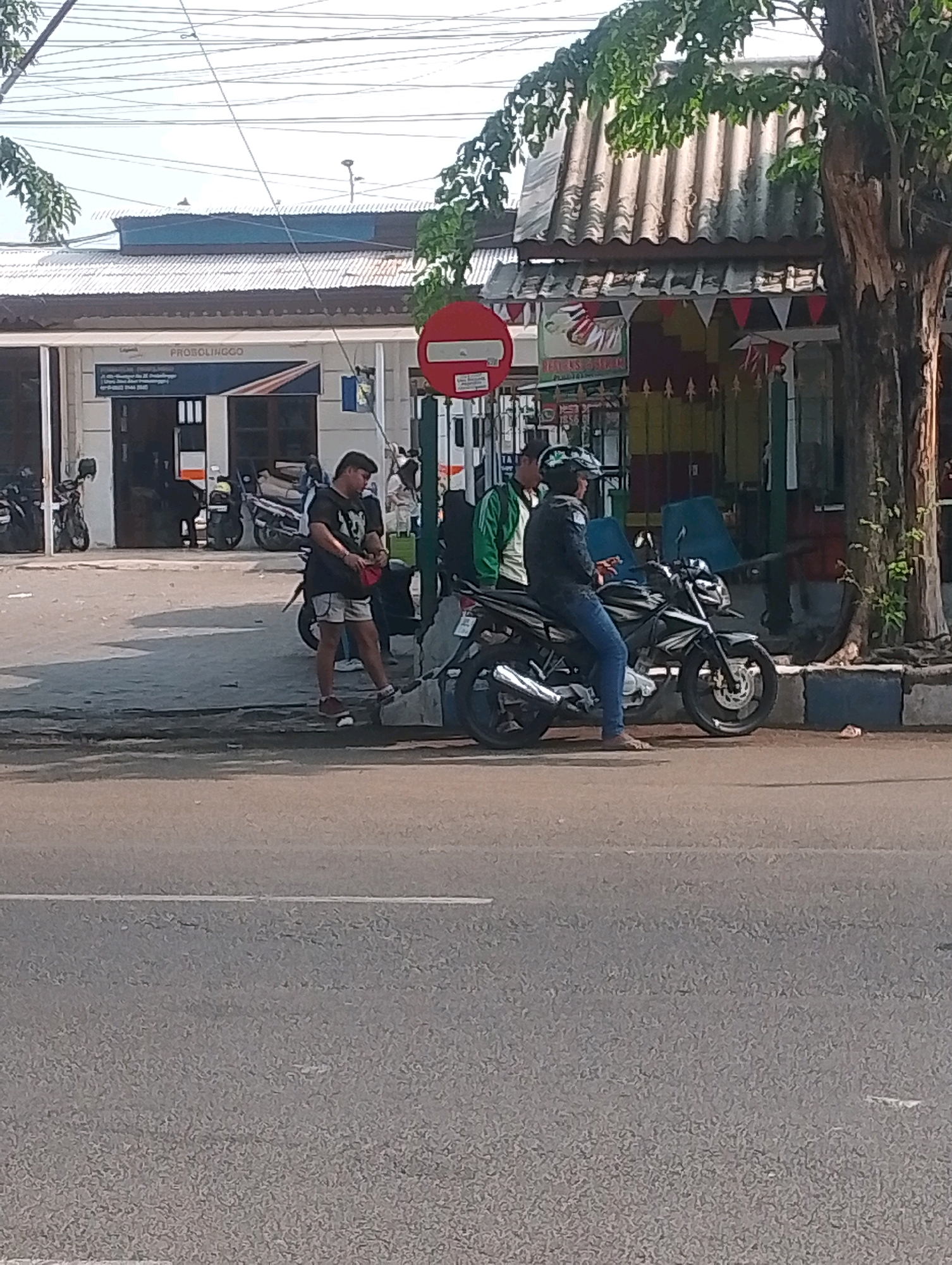 The image depicts a street scene in Probolinggo, Indonesia. In the foreground, a gray asphalt road is visible with a white line marking the edge. Behind the road, a paved sidewalk runs along the front of a building. Several people are present. A man in a black t-shirt and shorts stands on the sidewalk, holding a red object and a dark bag. To his right, two individuals are seated on a black motorcycle. One, wearing a helmet and jeans, is operating the motorcycle. The other person, wearing a green jacket, stands beside them. The building behind the sidewalk appears to be a commercial establishment with a corrugated metal roof. A red circular traffic sign with a white horizontal bar is positioned on the sidewalk. Several other motorcycles are parked in front of the building, and a large tree with rough bark is visible to the right of the scene. The sky above is clear and bright.
