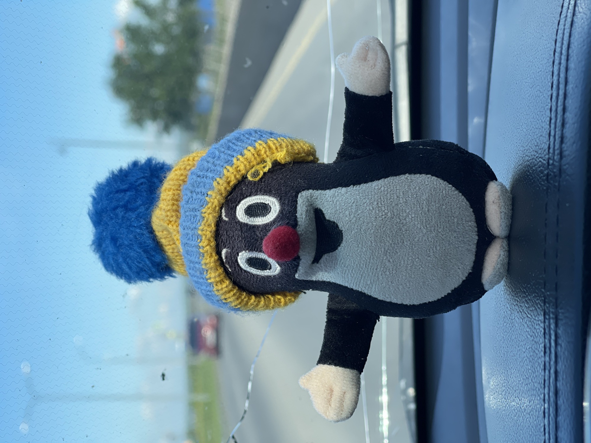 The image, originally rotated 90 degrees clockwise, prominently features a plush toy resembling Krtek, the Little Mole, situated indoors and facing outwards through a window. The toy has a dark brown or black body with a light grey belly, white eyes with black pupils, and a distinctive red nose. It is adorned with a knitted hat featuring stripes of light blue, yellow, and darker blue, topped with a fluffy blue pom-pom. Its small, rounded hands and feet are white.
The toy is positioned upright against a clear window. To its immediate right, a dark blue, textured surface, possibly a car seat or interior panel with visible stitching, forms part of the background. A thin, transparent string or wire is visible near the top of the toy, suggesting it might be suspended.
Through the window, a bright, clear blue sky occupies the upper background. The windowpane itself displays faint wavy distortions and minor smudges or dust. In the blurred distance, green foliage of trees and indistinct shapes, possibly other vehicles or structures, are visible, implying an open outdoor setting, likely from a moving or parked vehicle. The scene is brightly lit, indicating it is daytime with clear, sunny weather. There is no specific activity depicted, and no text is visible within the image.