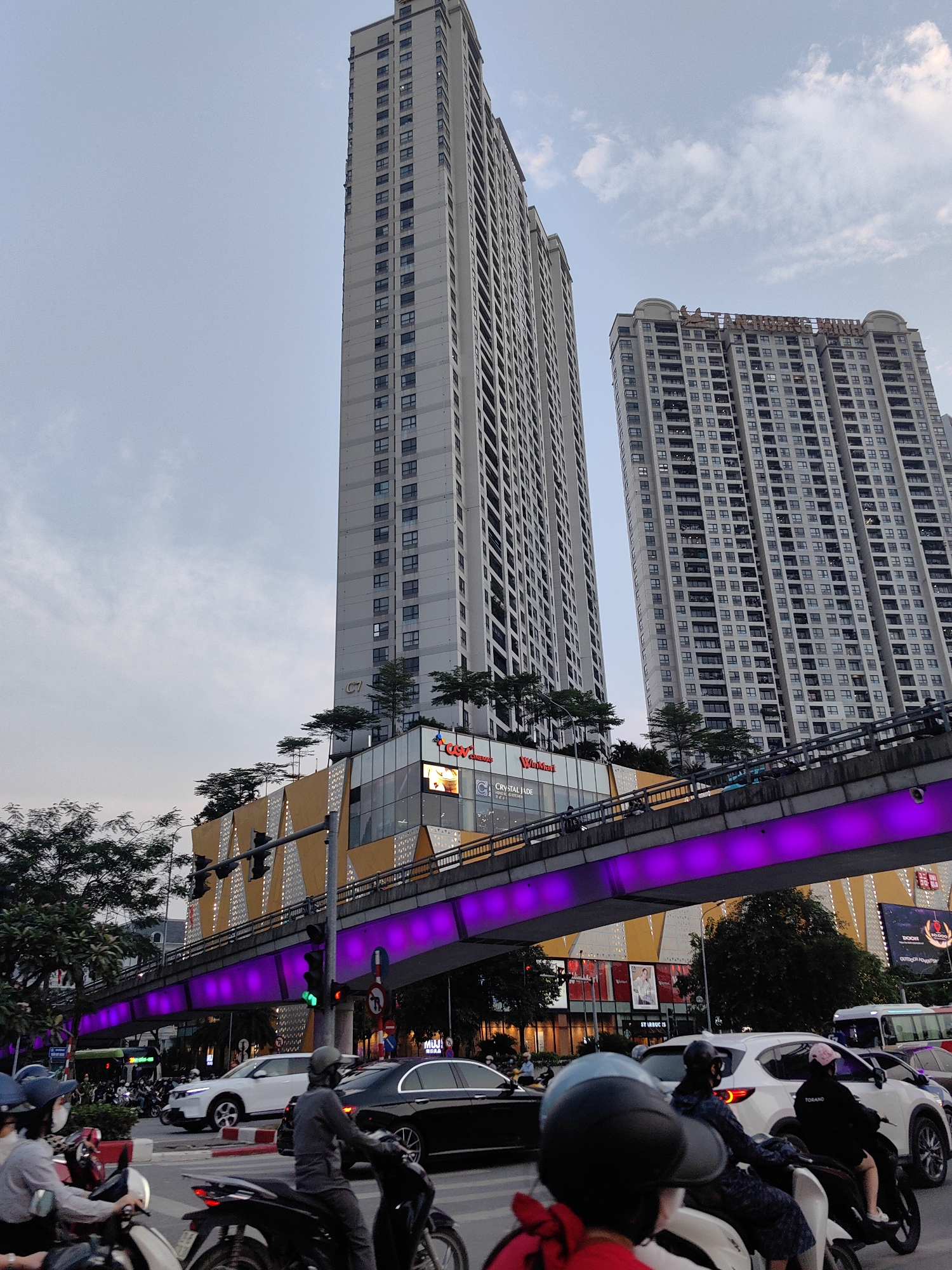 This image captures a vibrant urban scene in Hà Nội, Vietnam, during dusk or early evening. The view looks up towards two prominent, modern high-rise buildings that dominate the skyline, suggesting a dense commercial and residential area.
The taller building on the left features a multi-story commercial complex at its base, characterized by a striking golden-yellow facade with geometric patterns and multiple rooftop terraces adorned with small trees. Visible signs on this complex include "CGV Cinemas," "Welcom...", "Orange...", "CGI," "CRYSTAL JADE," and "STARBUCKS."
An elevated pedestrian bridge, brightly illuminated with striking purple LED lights, spans across the busy intersection below. This bridge serves as a prominent visual element, adding a modern, dynamic feel to the cityscape.
Below the bridge, the street is bustling with typical Hà Nội traffic. Numerous motorbikes, ridden by individuals wearing helmets, share the road with several cars, including a white SUV and a black sedan. A "TORANO" sticker is visible on the rear of a white car. A green traffic light indicates active vehicle movement.
On the right, another large high-rise building stands, with the word "VINHOMES" visible on its upper section. Its lower levels also appear to house commercial spaces, with partially visible signs for "UNIQLO," "MUJI," and "DOOM outdoor adventure." The sky is a soft blue-grey, indicating the transitional light conditions as day turns to night, with scattered clouds adding texture. The overall impression is one of a bustling, modern Vietnamese metropolis.