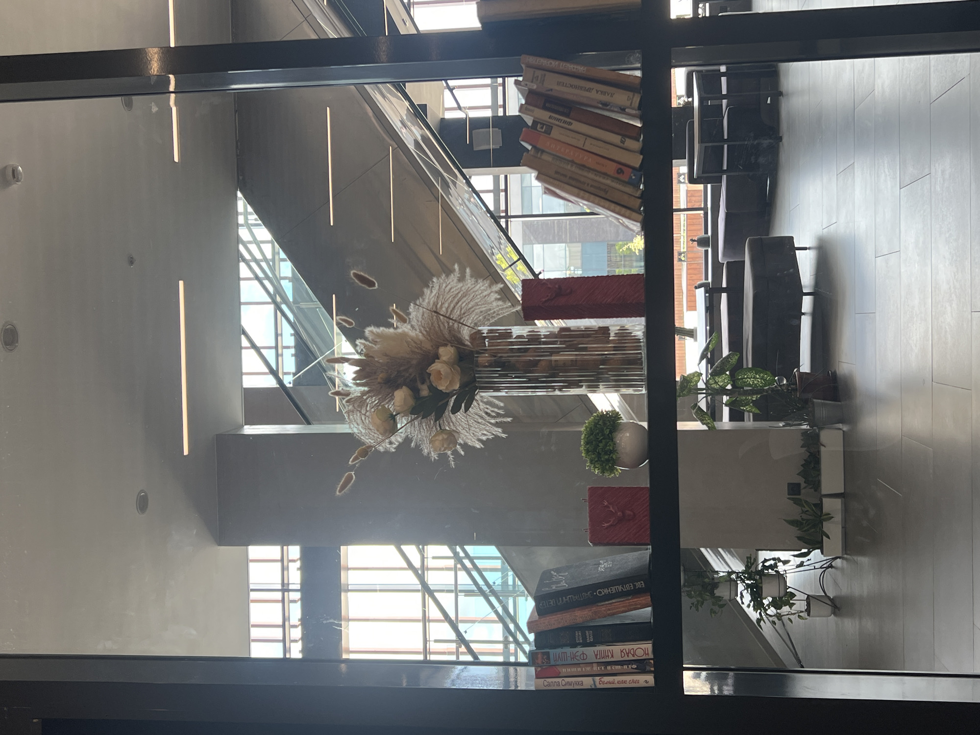 A shelf displays several books, a vase with dried flowers, a small potted plant, and two red decorative objects. The books are on the left side of the shelf, arranged in a row. The vase, filled with various dried floral elements, is positioned centrally behind the books. To the right of the vase, a small green potted plant sits in a white ceramic pot. Two rectangular red decorative items are placed to the right of the plant. The shelf is viewed through a glass pane, reflecting the interior of a modern building with escalators and seating areas. The exterior environment visible through the windows suggests a bright, possibly daytime setting with architectural structures. The scene appears to be located within a public space, likely a library, cafe, or office building. The specific location in Veliky Novgorod, Russia, cannot be determined from the visual information provided.