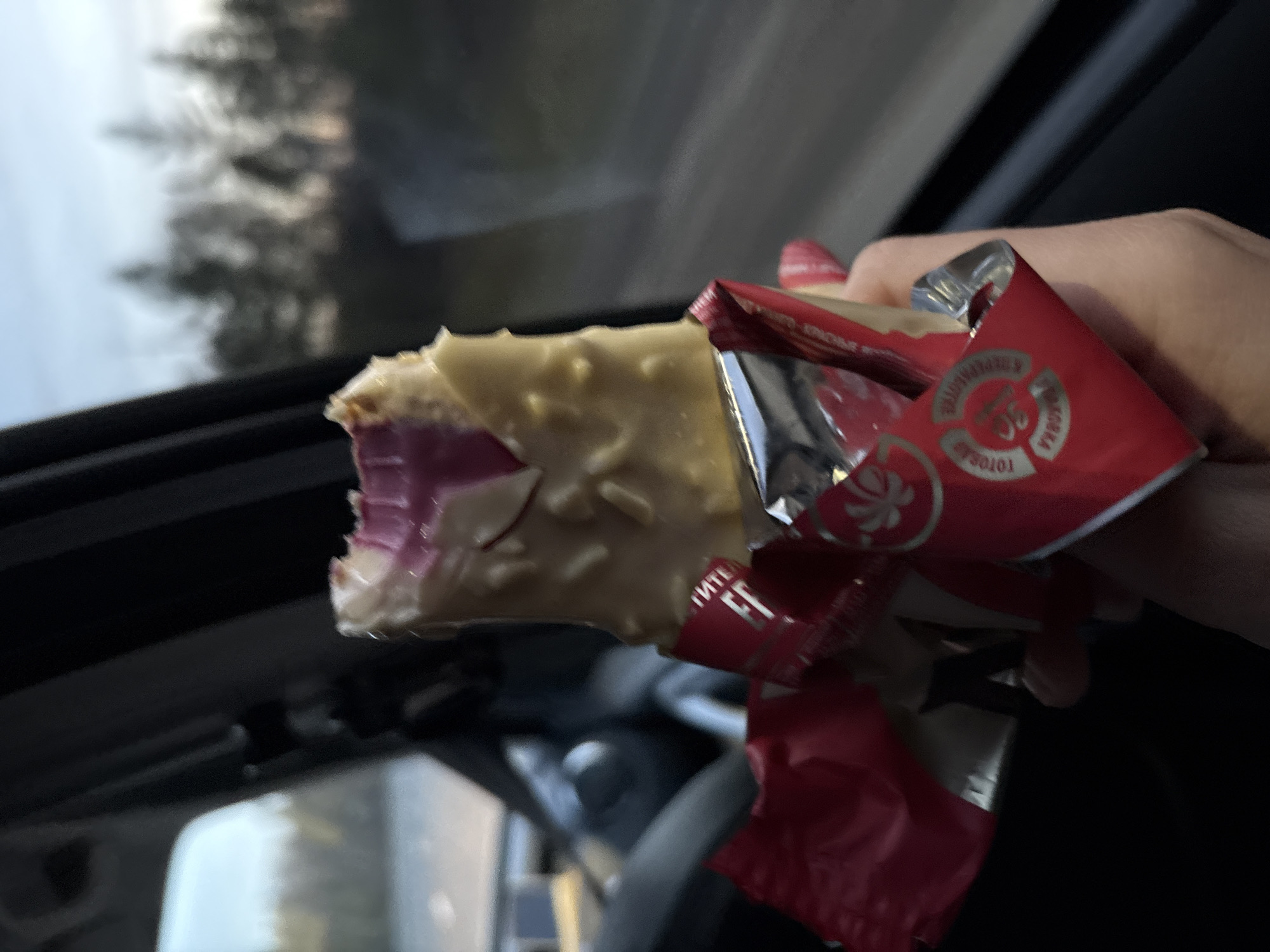 A close-up, slightly blurred image shows a person's hand holding a partially eaten ice cream bar in a car. The ice cream has a white coating with small pieces, a pink interior, and visible bite marks. The wrapper is red and silver, with Russian text and circular designs. The car interior is visible in the background, including parts of the steering wheel, dashboard, and the outdoors seen through the window, including the sky and trees. Based on the packaging, the image was likely taken in Russia. Given the information on the wrapper, the location may be Bratsk, Russia.