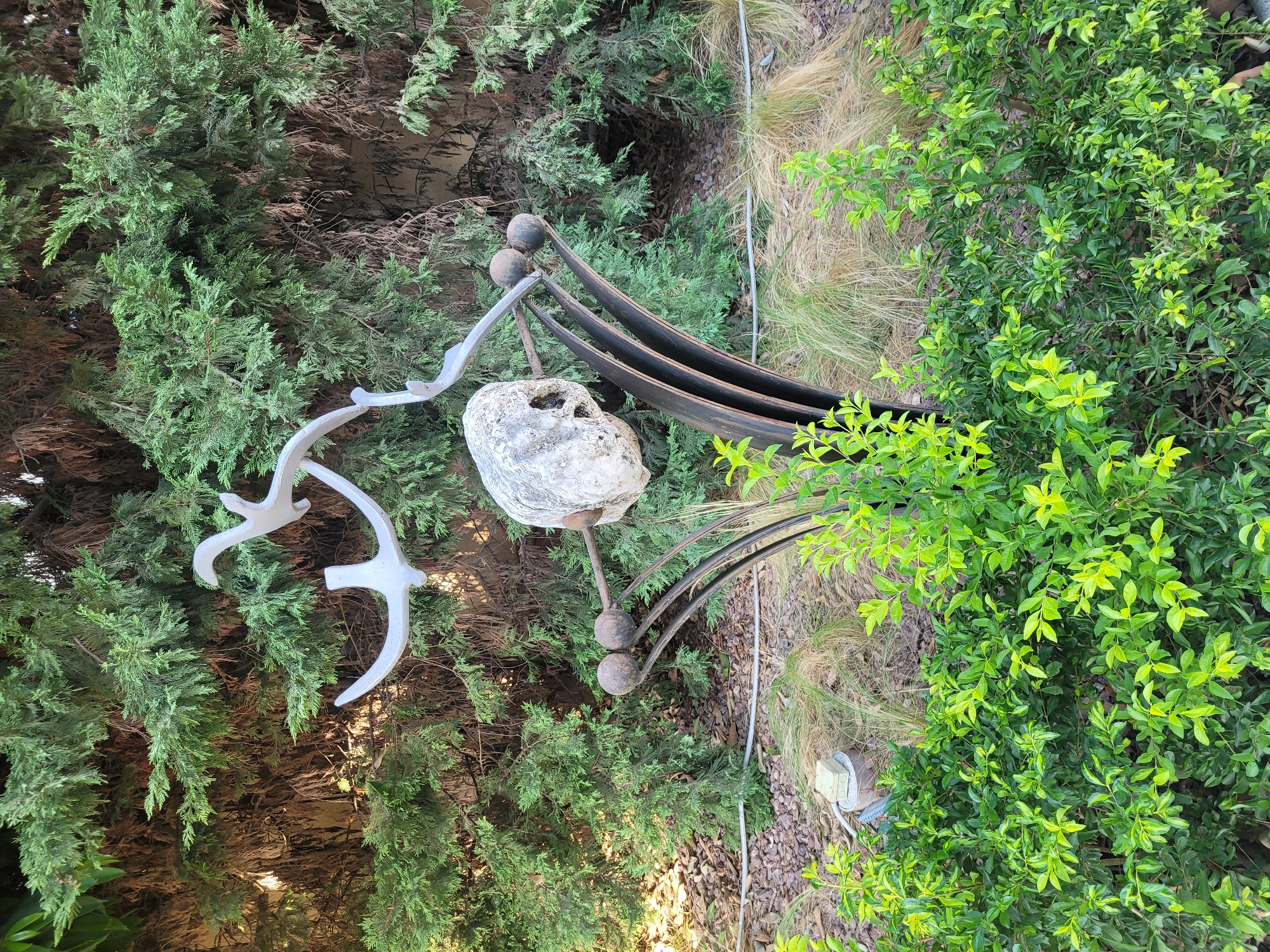 The image shows an outdoor sculpture featuring a white metal bird motif with two abstract bird figures in flight, mounted on a dark metal base. The base is characterized by curved, layered elements and a rough, light-colored stone positioned centrally. The sculpture is situated in a garden environment with lush green bushes in the foreground and a dense, dark green evergreen tree line in the background. There are no people or animals present in the image. The overall scene appears to be a quiet, possibly artistic, outdoor setting. This sculpture is located in Limassol, Cyprus, specifically in the Municipal Gardens.