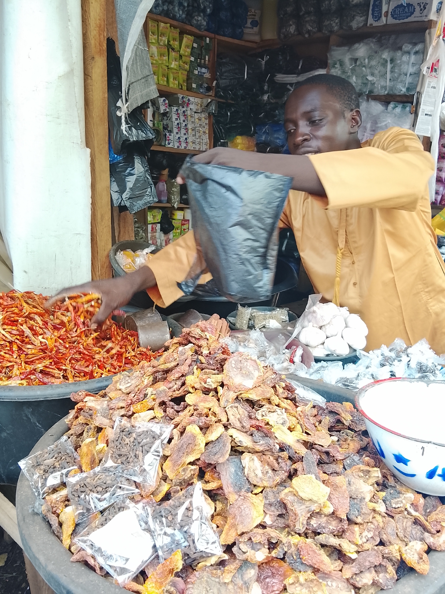 In a market stall, a young Black man wearing a bright yellow robe stands behind a display of various dried goods. His right hand is outstretched, holding a black plastic bag over a large, dark gray basin overflowing with bright red dried chili peppers. In front of the chilies, another large basin is filled with a mix of dried goods, predominantly brown and orange pieces that appear to be dried fruits or vegetables, along with several small, clear plastic bags containing dark brown, small-grained spices. To the right of these basins, a pile of white garlic bulbs is visible, some individually wrapped in plastic. Further to the right, a white enamel bowl with a blue and red rim is partially visible, filled with a white substance, possibly salt or sugar. The stall is lined with shelves in the background, stocked with various packaged goods, including what appears to be small bags of spices or food items. The overall impression is a bustling marketplace scene.