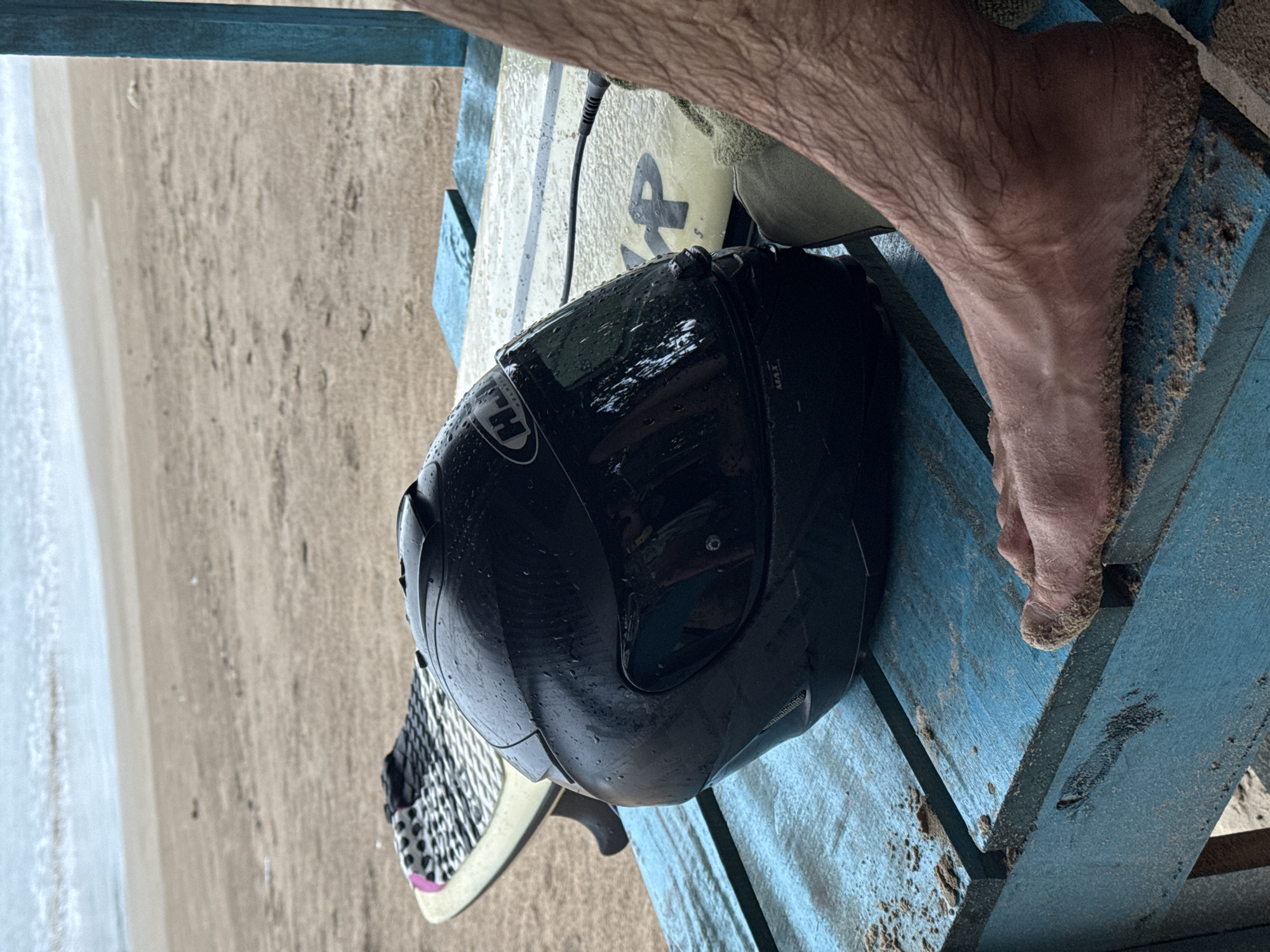 A person's sandy foot rests on a blue wooden bench, next to a black helmet and a surfboard. The scene appears to be on a beach, with sand and the ocean visible in the background. The helmet is wet, suggesting recent activity or rain. The surfboard has some writing on it, including the number "4". The overall impression is of a casual, outdoor setting, likely in a tropical location given the sand and apparent warmth. The image conveys a sense of relaxation and leisure, possibly after a day of surfing or water sports. The time of day is not explicitly indicated, but the lighting suggests it could be daytime. The presence of sand on the bench and foot indicates the proximity to the beach.