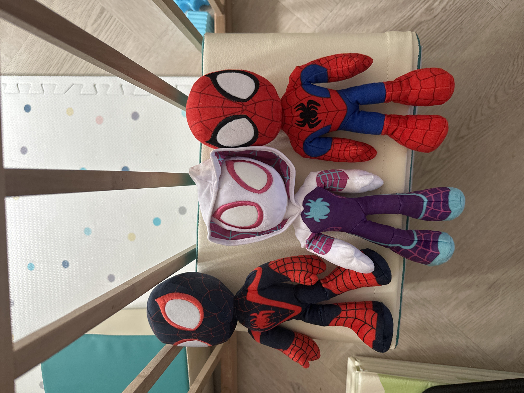 Three Spider-Man plush toys are arranged in a line, standing on a beige cushioned surface. The toy on the left is Miles Morales, dressed in a black and red suit with a spider emblem on his chest. The middle toy is Spider-Gwen, wearing a white hooded suit with purple accents and a spider emblem. The toy on the right is the classic Spider-Man, in his red and blue suit with a spider emblem. They are positioned in front of a light-colored mat with colorful polka dots. The scene appears to be indoors, possibly a playroom or nursery, with wooden bars and a light-colored floor visible. There is no indication of the location being in Leeds, United Kingdom.