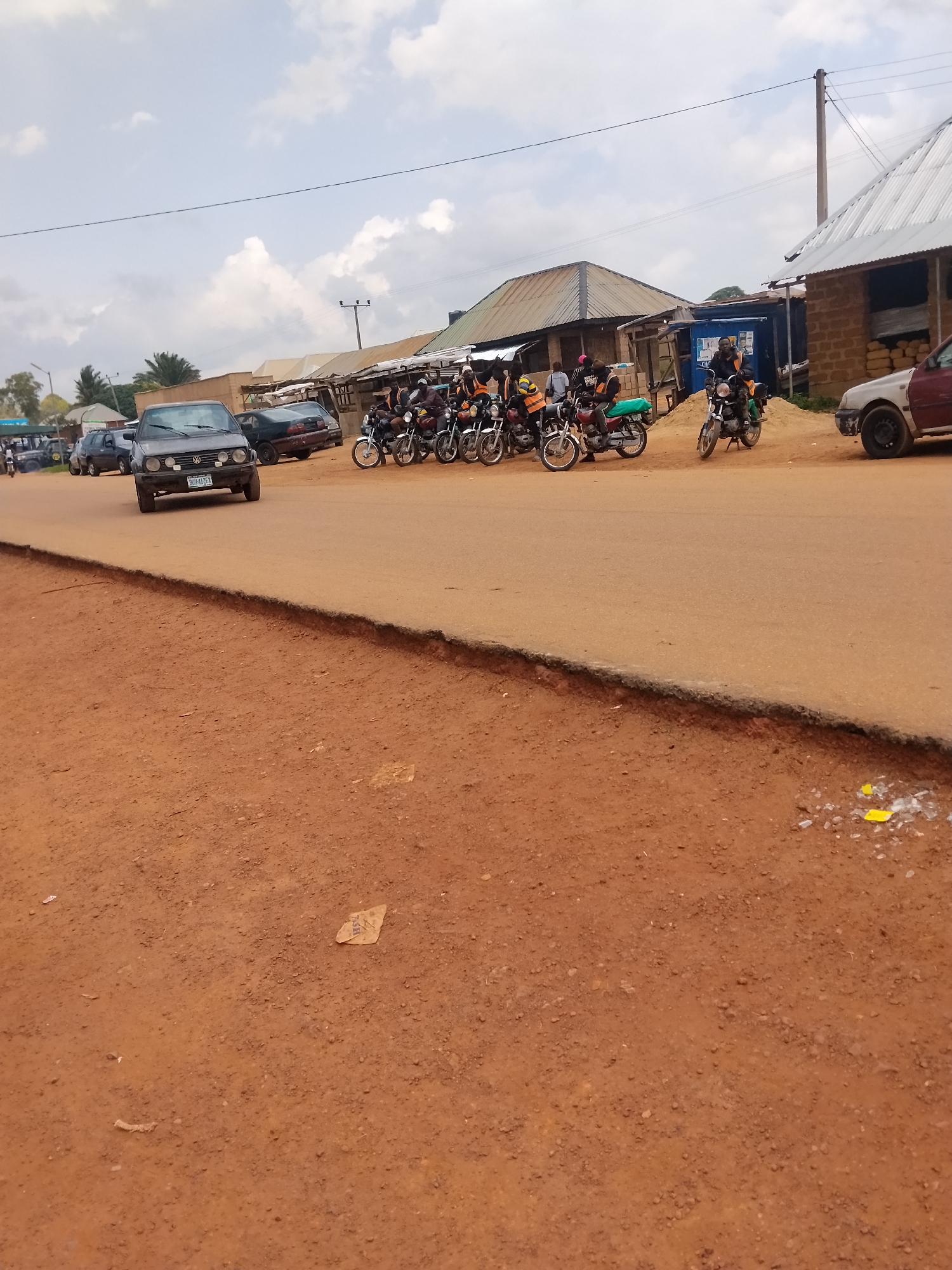 The image depicts a street scene in Durumi, Nigeria, on an overcast day. A red dirt road dominates the foreground, with a paved road to the left where several vehicles are present. A dark grey Volkswagen Golf is in the center-left, facing forward, its license plate reading "BDU 417EX". To its right are two other cars, one black and one dark blue. A line of motorcycles, mostly with riders, is parked on the right side of the paved road. The motorcycles are of various colors, including red and black. Several individuals are seen on or around the motorcycles, some wearing orange vests, suggesting they might be commercial motorcycle riders, often referred to as "Okada" in Nigeria. In the background, buildings with corrugated metal roofs are visible, typical of urban or semi-urban areas in Nigeria. The sky is cloudy, with patches of blue peeking through. The overall impression is of a busy, albeit dusty, street, likely a commercial hub or a common meeting point for transportation.
