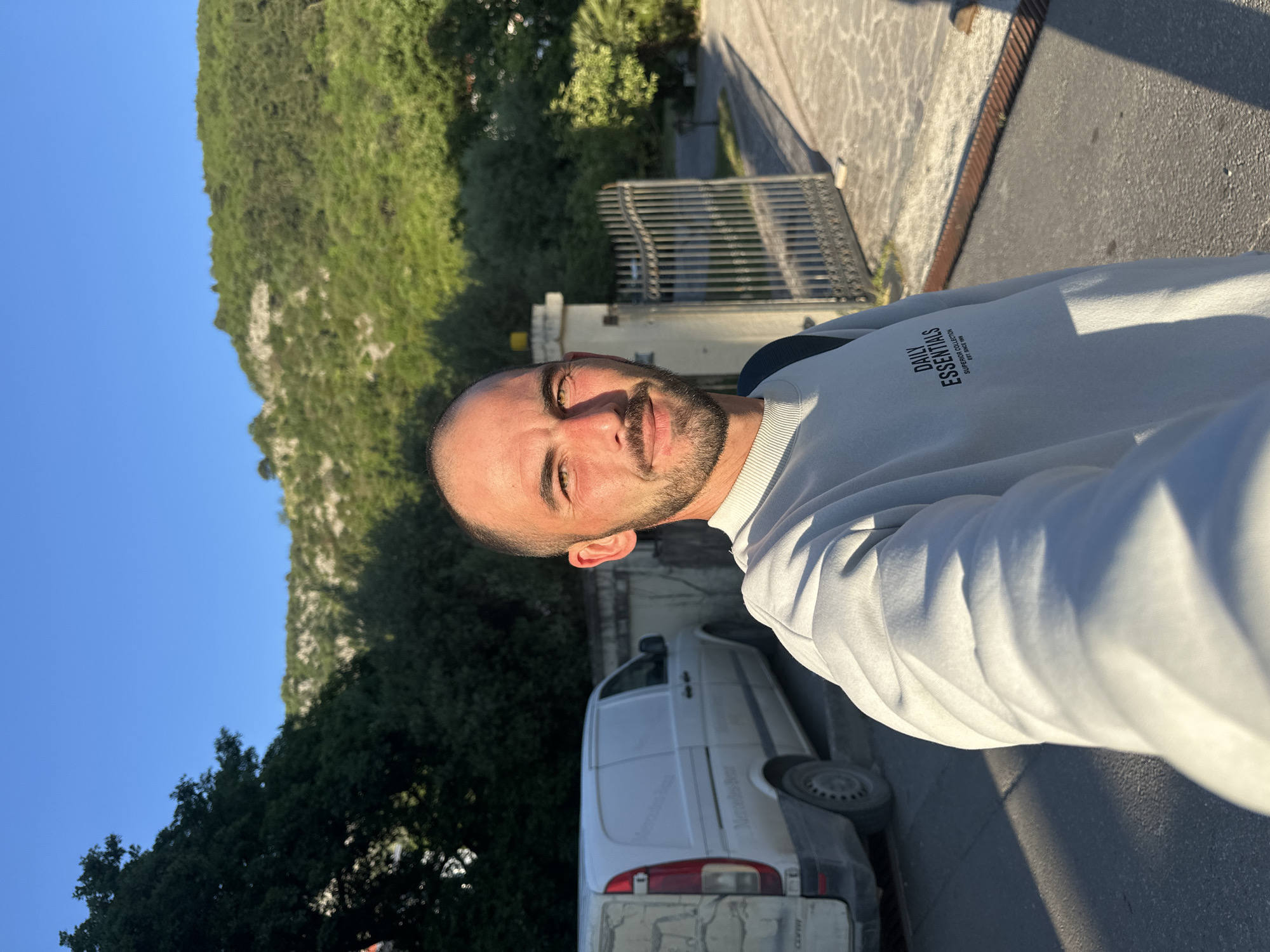 A selfie shows a man with short dark hair and a beard. He wears a light-colored sweatshirt with the words "DAILY ESSENTIALS SUPERIOR COLLECTION EST. SINCE 1996".
Behind the man are green trees and a hillside under a blue sky, likely a mountain slope. A white van is parked nearby, and there is a metal gate and concrete pathway.
This photo was taken in Budva, Montenegro.