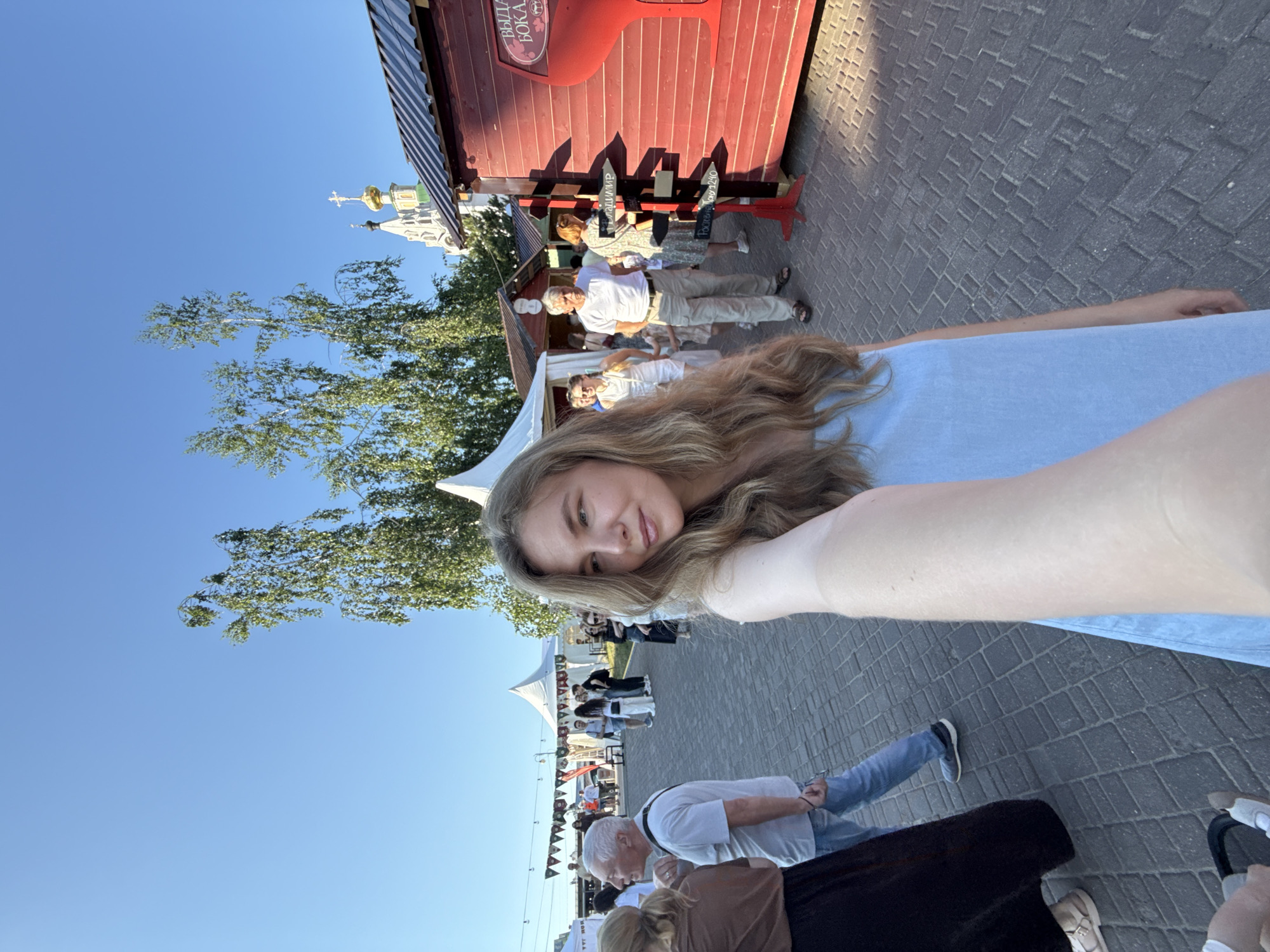 A woman in a light blue dress takes a selfie at an outdoor market in Vladimir, Russia. She stands in the foreground, looking to her left. Behind her, a crowd of people walks through stalls and tents, some with white conical roofs. To her right, a red wooden building with a sign that reads "ВЫДАЧА БОКАЛ" (Vydacha Bokala) and a large wine glass graphic is visible. A wooden directional sign with arrows pointing to "Владимир" (Vladimir) and "Ростов (на Дону) 1240" (Rostov (on Don) 1240) stands in front of the red building. In the background, trees and the golden domes of a church can be seen against a clear blue sky. The ground is paved with dark grey cobblestones.