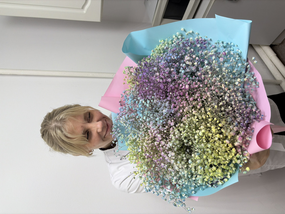 The image displays a blonde female in an indoor setting, holding a large, multi-colored bouquet of dyed Gypsophila. The woman, appearing to be in her 30s or 40s, wears a white uniform-style top and smiles gently while directing her gaze towards the flowers. The spherical bouquet is composed of distinct sections of yellow, green, blue, purple, and pink baby's breath, and is wrapped in an outer layer of light blue paper over an inner layer of light pink paper. The background consists of plain white walls. A white vertical pipe runs along the wall to the right of the subject, adjacent to partially visible white cabinetry and a dark appliance, possibly a microwave, on a counter. The specific location within Chaikovsky, Russia, cannot be determined from the visual information provided.