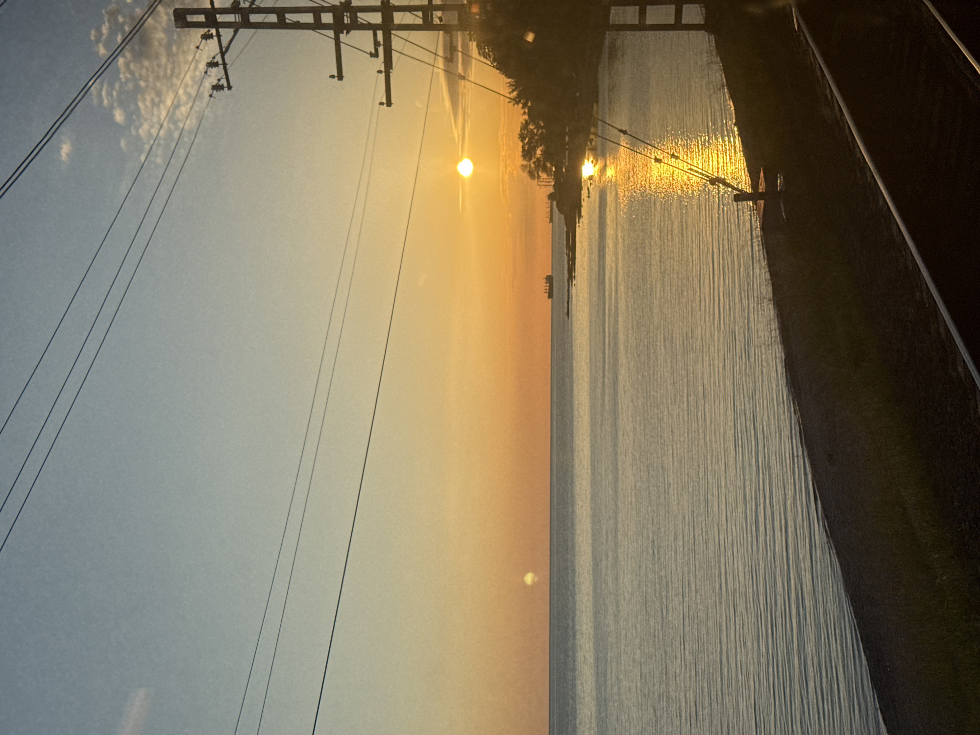 The image, taken in what appears to be Vishnevka, Russia, captures a serene sunrise or sunset over a body of water. The scene is dominated by the warm, golden light of the low-lying sun on the horizon, which casts a brilliant, shimmering path across the calm water surface.
The sky is a gradient of colors, transitioning from a light blue with a few wispy clouds in the upper portion to a vibrant golden-orange closer to the horizon where the sun is visible. Multiple dark power lines stretch diagonally across the upper left and center of the frame, supported by utility poles that are partially visible in silhouette.
A dark, silhouetted landmass, possibly a shoreline or embankment densely covered with trees, separates the power lines from the expanse of water. On the far right, a dark, angled structure, which could be a road or a concrete barrier, runs parallel to the water's edge. Faint, dark shapes, possibly distant boats or structures, are visible along the horizon within the sun's reflection on the water.
No people are present in the image, emphasizing the natural beauty and tranquility of the moment. The calm water, soft light, and absence of activity evoke a peaceful atmosphere. There is no visible text.