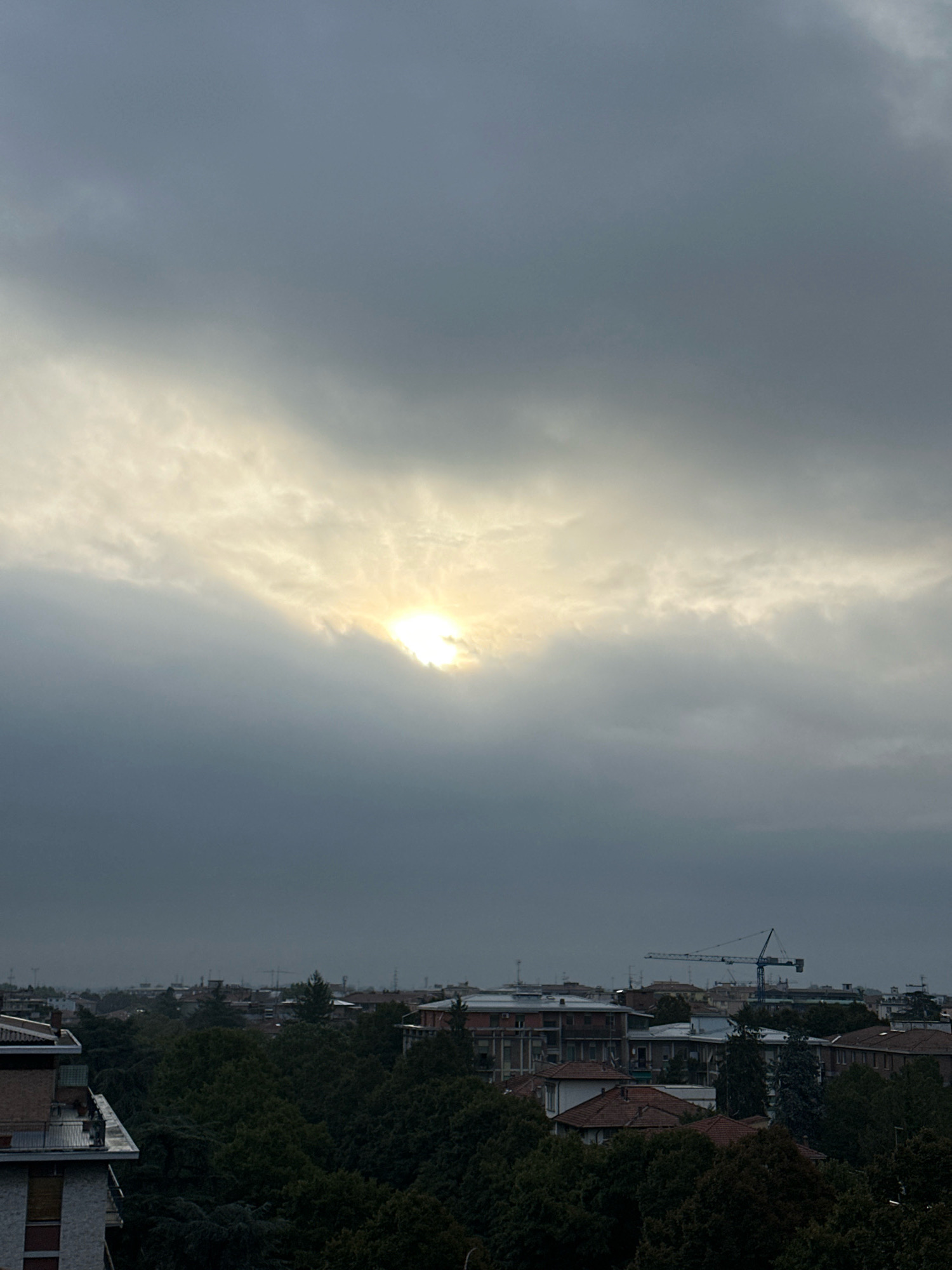 This image captures a cloudy sky over the city of Parma, Italy, with a sliver of the sun breaking through the clouds in the upper center. The foreground features a cluster of buildings and lush green trees. In the middle ground, more buildings and trees are visible, with a prominent construction crane situated among them. The overall atmosphere is subdued due to the overcast conditions, suggesting it might be early morning, late afternoon, or a day with poor weather.