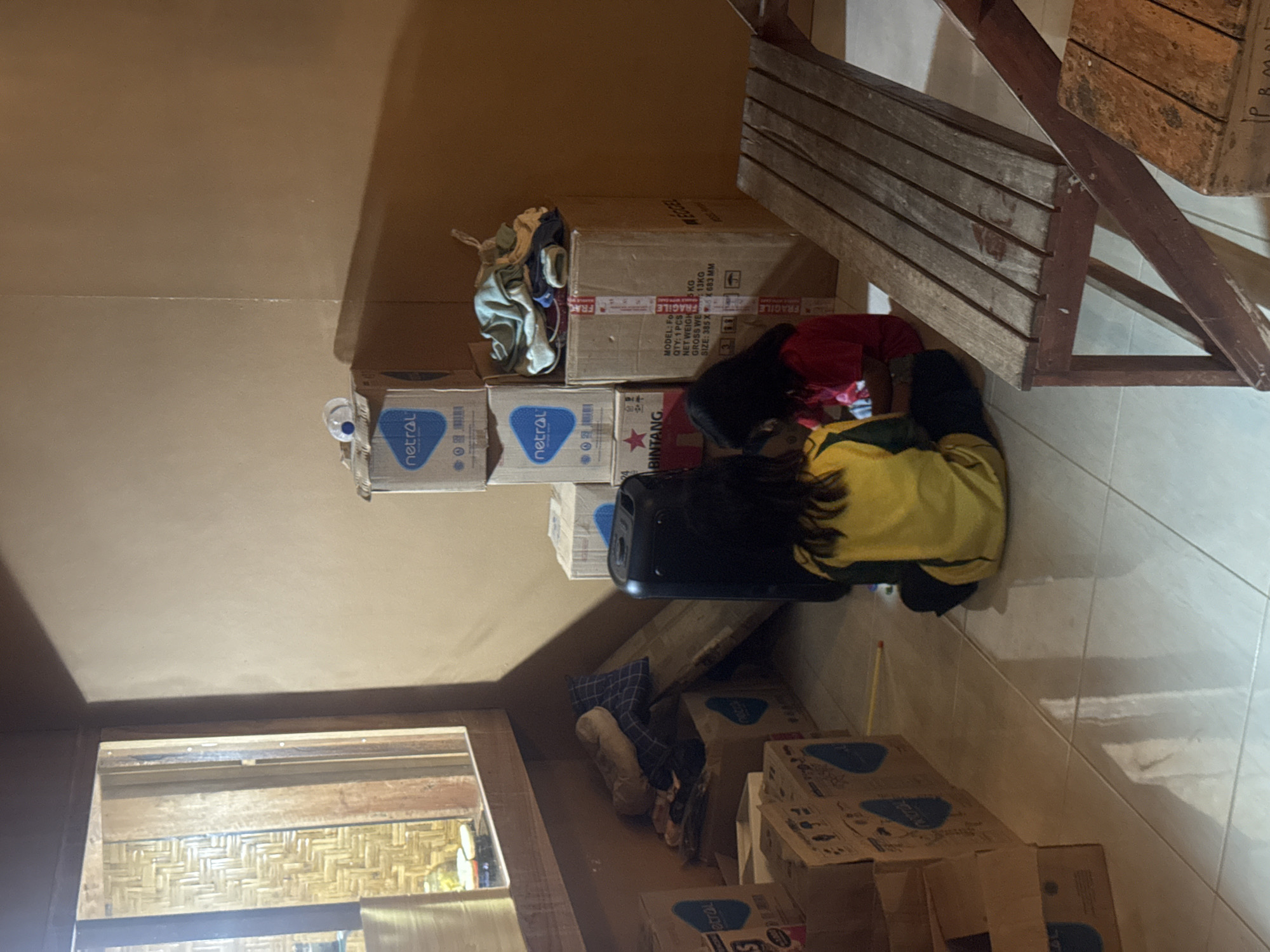 In the city of Kertasari, Indonesia, an indoor scene is depicted, likely a storage area or room. Two young girls, one wearing a yellow and green shirt and the other a red shirt, are seated on the floor amongst stacks of cardboard boxes. The boxes, some bearing the "netral" logo and "BINTANG" branding, suggest potential goods for storage or transport. A large black speaker or electronic device stands to the left of the girls. A wooden bench is positioned in the foreground, to the right, with an open wooden crate beside it. The walls are a warm, earthy tone, and the floor is tiled. Natural light filters in from a window on the left, revealing a woven bamboo texture behind a wooden frame. The overall impression is one of a temporary storage space or a home environment with unpacked or stored items.
