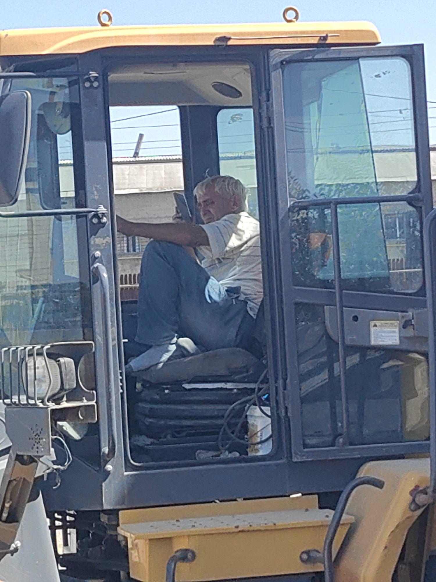 A man with gray hair is sitting in the cab of a yellow construction vehicle, possibly a backhoe. He is wearing a light-colored striped shirt and blue jeans. His right arm is resting on the door frame, and he appears to be holding a dark-colored object, possibly a cell phone, in his hand. The cab has large glass windows. The vehicle is parked outdoors, with buildings and power lines visible in the background. The sky is clear and blue. The image was taken in Shymkent, Kazakhstan.