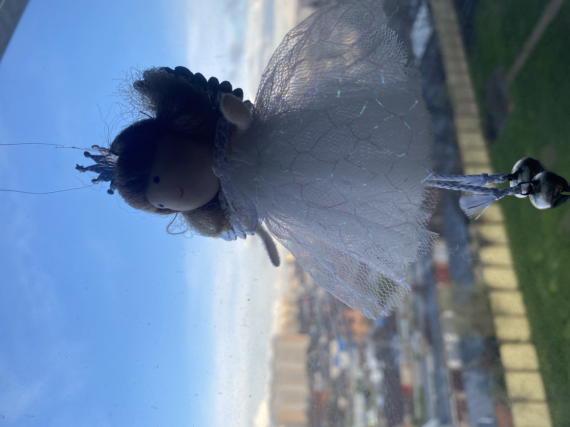 A close-up shot of an angel ornament hanging in front of a window. The angel has a beige face, brown hair, and a white tulle dress. Behind the angel, the window offers a view of a city skyline with various buildings and greenery visible. The sky is blue with some clouds. The location appears to be a high-rise building in an urban area.