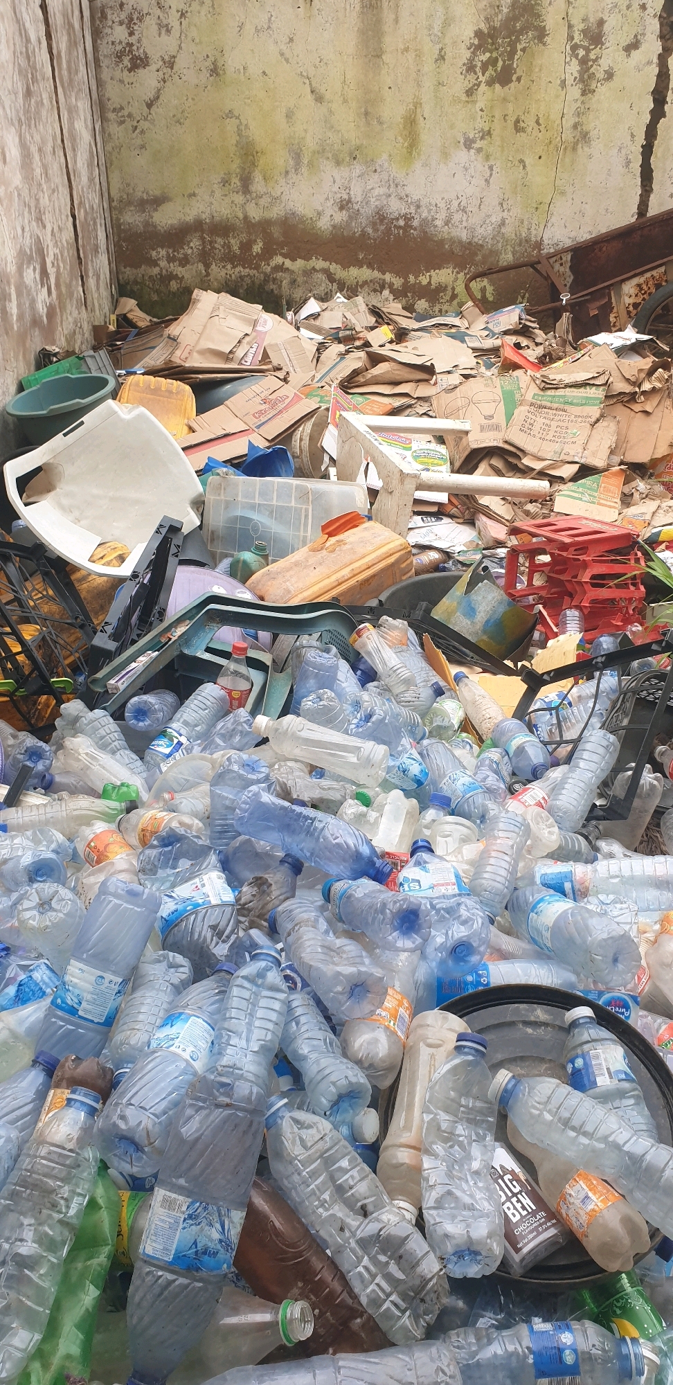 A pile of recyclable waste is located in a concreted corner of a property in Sabon Tasha, Nigeria. The waste consists primarily of plastic bottles, flattened cardboard boxes, and some miscellaneous plastic containers and buckets. A large, dirty, off-white wall forms the backdrop, with visible signs of dampness and algae growth. The ground is covered in a thick layer of debris. Among the scattered items, a rusty wheelbarrow is partially visible. The overall scene depicts a disorganized accumulation of discarded materials.