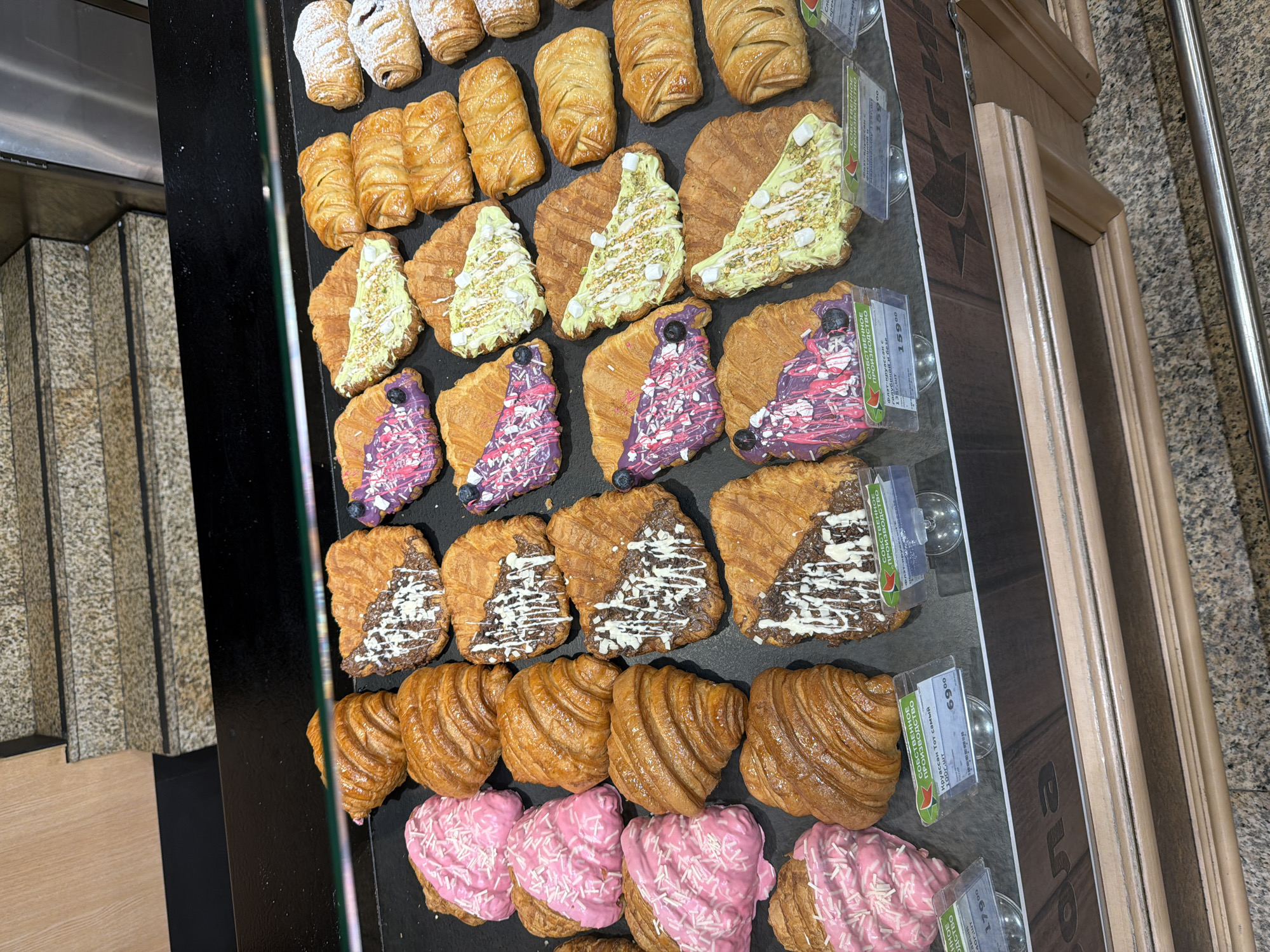 The image shows a display case of pastries in what appears to be a bakery or food establishment in Yekaterinburg, Russia. There are several types of pastries including croissants with various toppings such as pistachio cream, purple icing with sprinkles, chocolate and white icing, and pink icing. Price tags in Russian are visible beside some of the pastries. The pastries are arranged in rows. To the left of the display case is a tiled wall. To the right is a wooden structure.