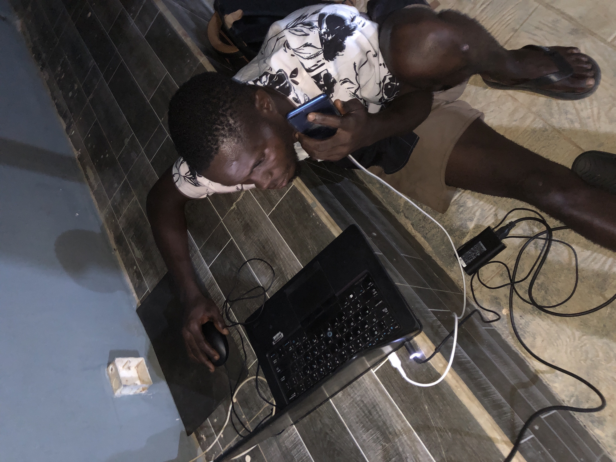 An overhead shot depicts a dark-skinned man lying on his stomach on a tiled floor, with his head near a laptop. He is holding a smartphone to his ear with his right hand, while his left hand is on a computer mouse. The man is wearing a light-colored shirt with a dark pattern and khaki shorts. The black laptop is connected to multiple cords, including a charging cable and a mouse. One leg is extended to the right, wearing a flip-flop. A white electrical outlet is visible on the wall near the laptop. The location is in the city of Unknown, Unknown.