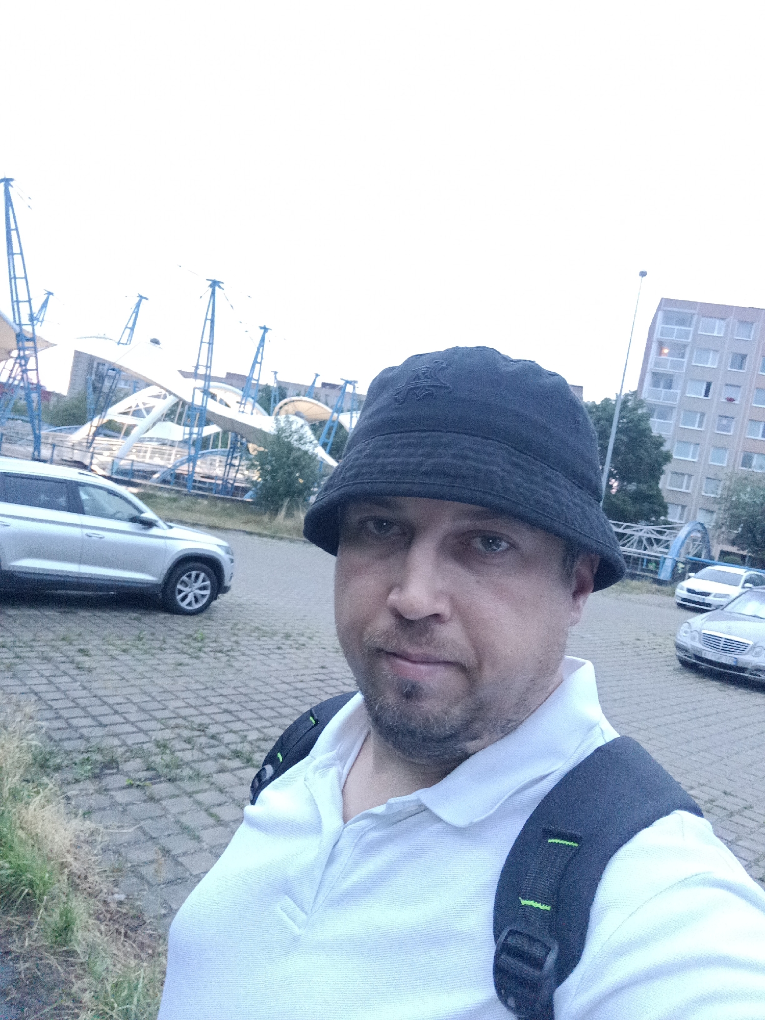 A selfie captures a man wearing a black bucket hat and a white polo shirt, with a black backpack on. He stands outdoors in what appears to be a parking lot with a patterned pavement. In the background, a silver car is parked to the left. Further back, there is an architectural structure with white and blue elements, possibly a stadium or a large canopy. To the right, a multi-story building and a street lamp are visible, along with two cars parked further down the lot. The scene is set during the day, under an overcast sky. Based on the architectural style of the building and the general urban environment, this location could be in Prague, Czechia, possibly near a public transportation hub or a recreational area.