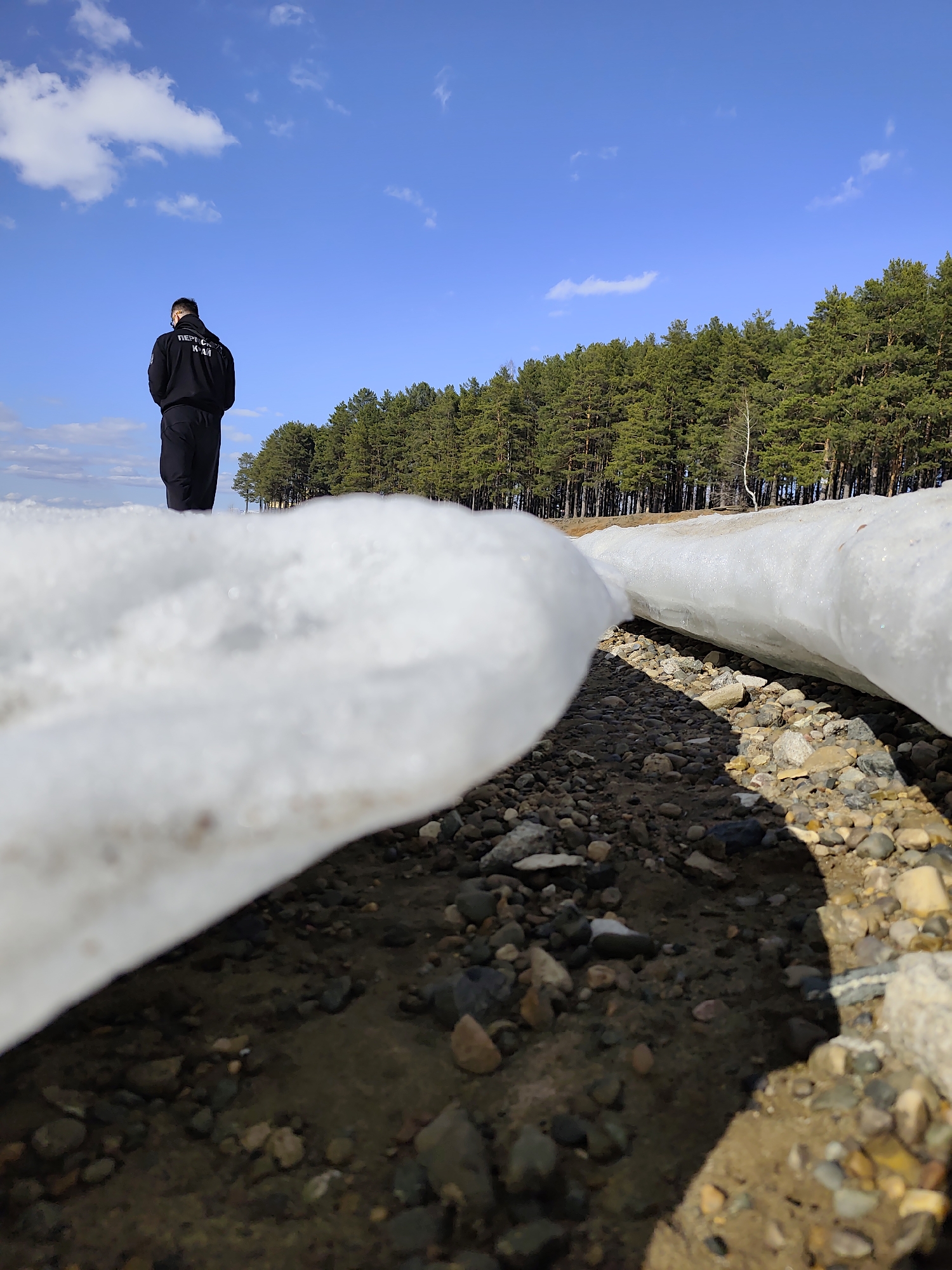 A low-angle, eye-level shot depicts a person standing on a snow-covered or icy shoreline, facing away from the camera, with a forest and blue sky in the background. The person wears a dark jacket with Cyrillic lettering and dark pants. In the foreground are smooth, water-worn pebbles and two large chunks of melting ice. The location is Chaikovsky, Russia.