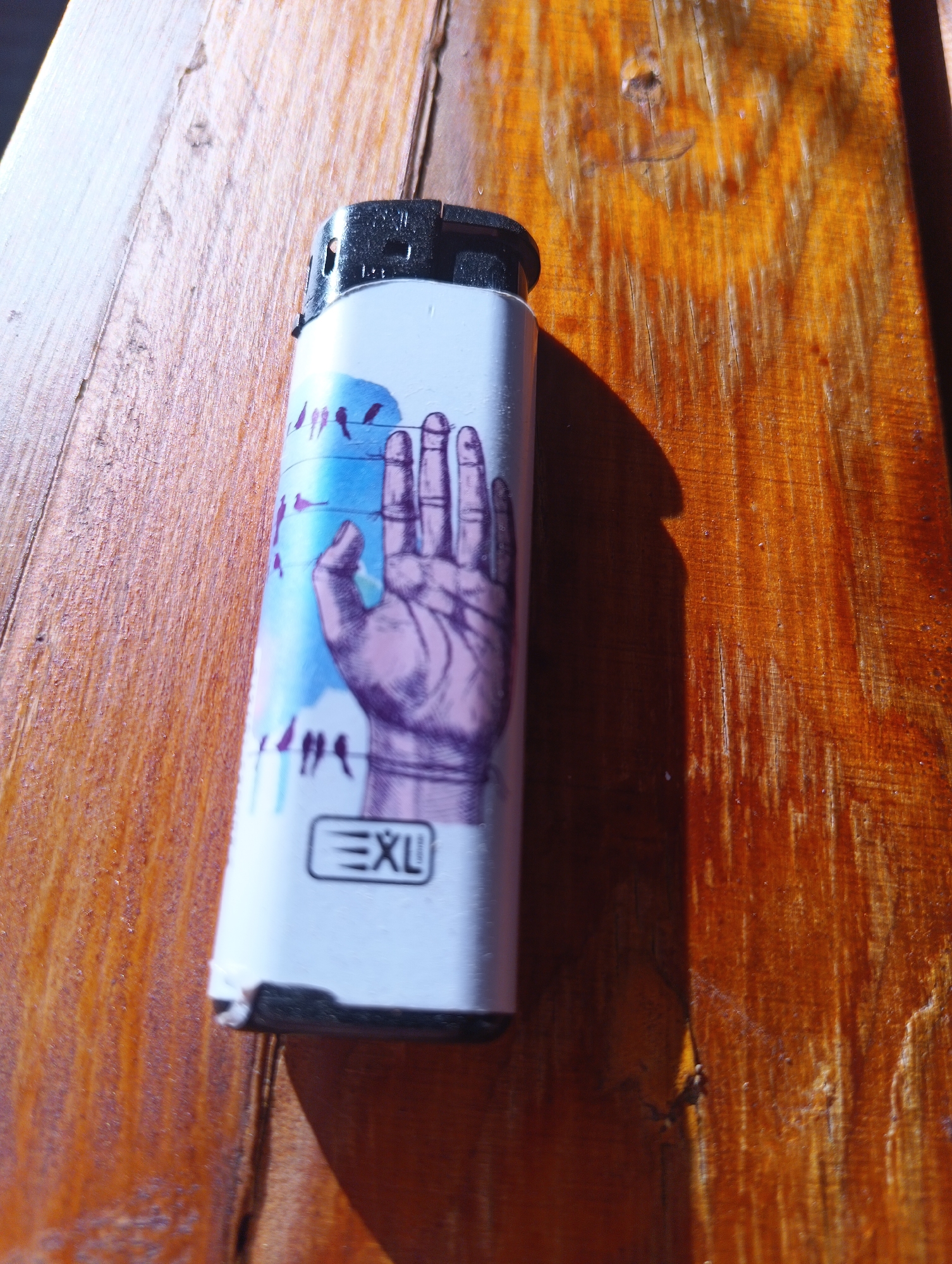 A close-up shot shows a white lighter with a graphic of a hand and birds on it, resting on a wooden surface. The lighter is positioned diagonally across the frame, with its black cap at the top left. The wooden surface appears to be weathered and textured, with visible grain and some imperfections. The background is a wooden table with warm, reddish-brown tones, illuminated by sunlight which casts a distinct shadow of the lighter. The scene is located outdoors on a wooden table in the city of Karnobat, Bulgaria.