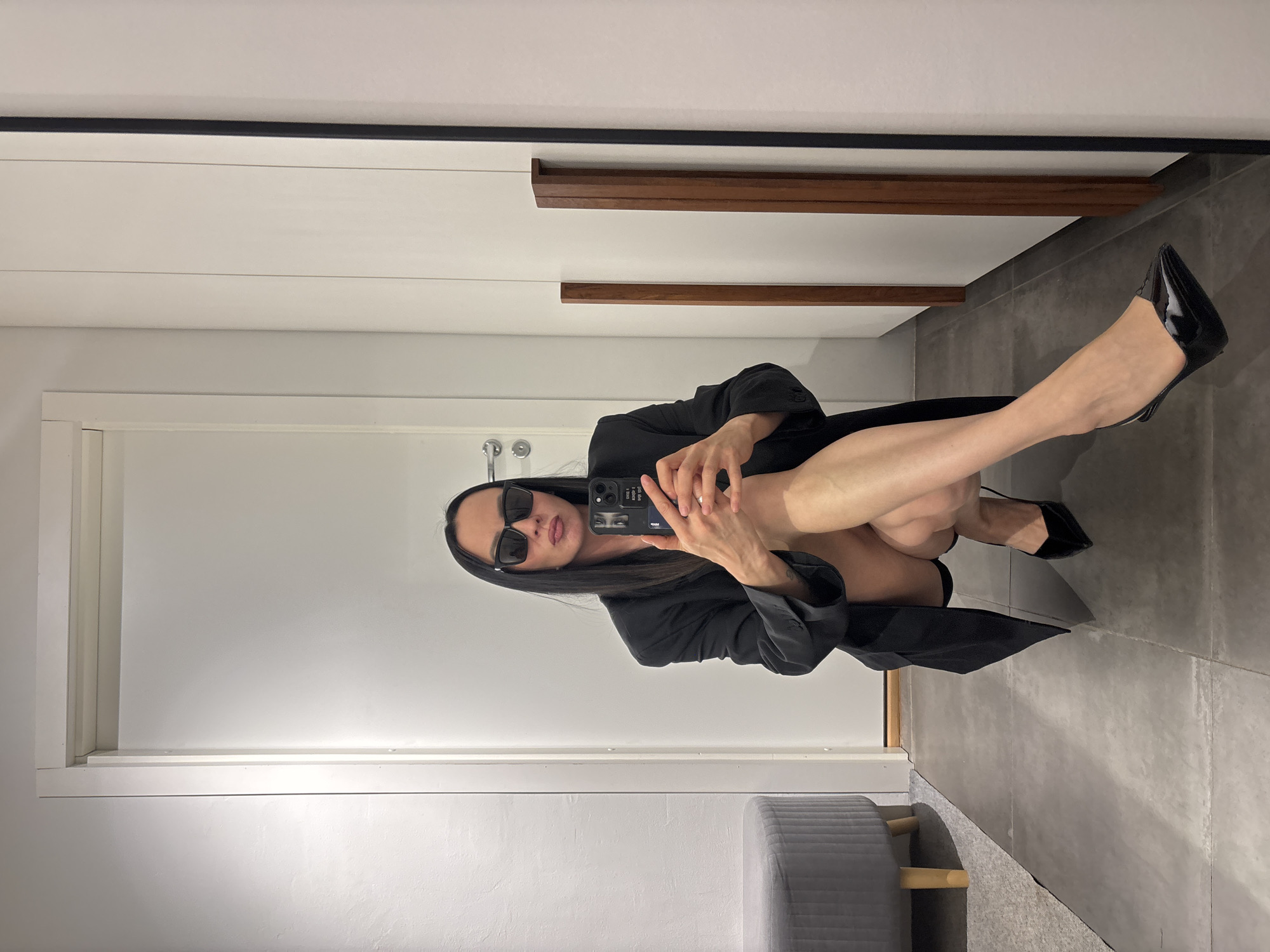 A woman wearing sunglasses and a black blazer is crouching and taking a selfie. She is also wearing black high heels. She is in a room that appears to be a dressing room or bathroom, with white walls and gray tile flooring. There is a gray ottoman in the corner. A partially visible door, a wooden ledge, and trim are also present. The image was taken in Chaikovsky, Russia.