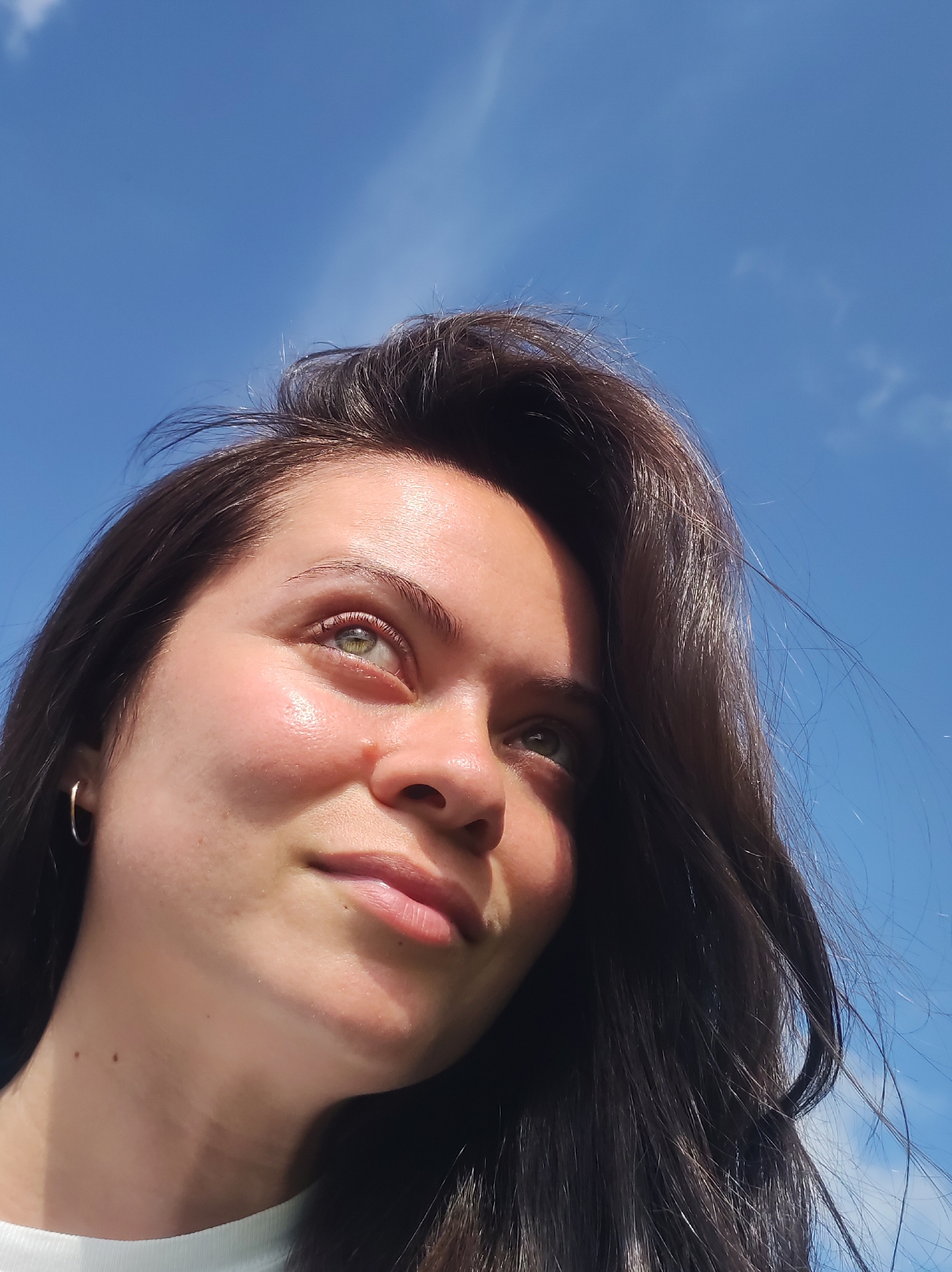 A young woman with long, dark hair and green eyes is pictured outdoors against a bright blue sky with wispy clouds. She is looking upwards and to the right, with a slight smile. Her left cheek is illuminated by sunlight, highlighting a faint sheen of sweat on her skin. She is wearing a small hoop earring in her left ear. The bottom of her neck and a portion of her white shirt are visible. There is no indication of the city of Foky, Russia, in the image.
