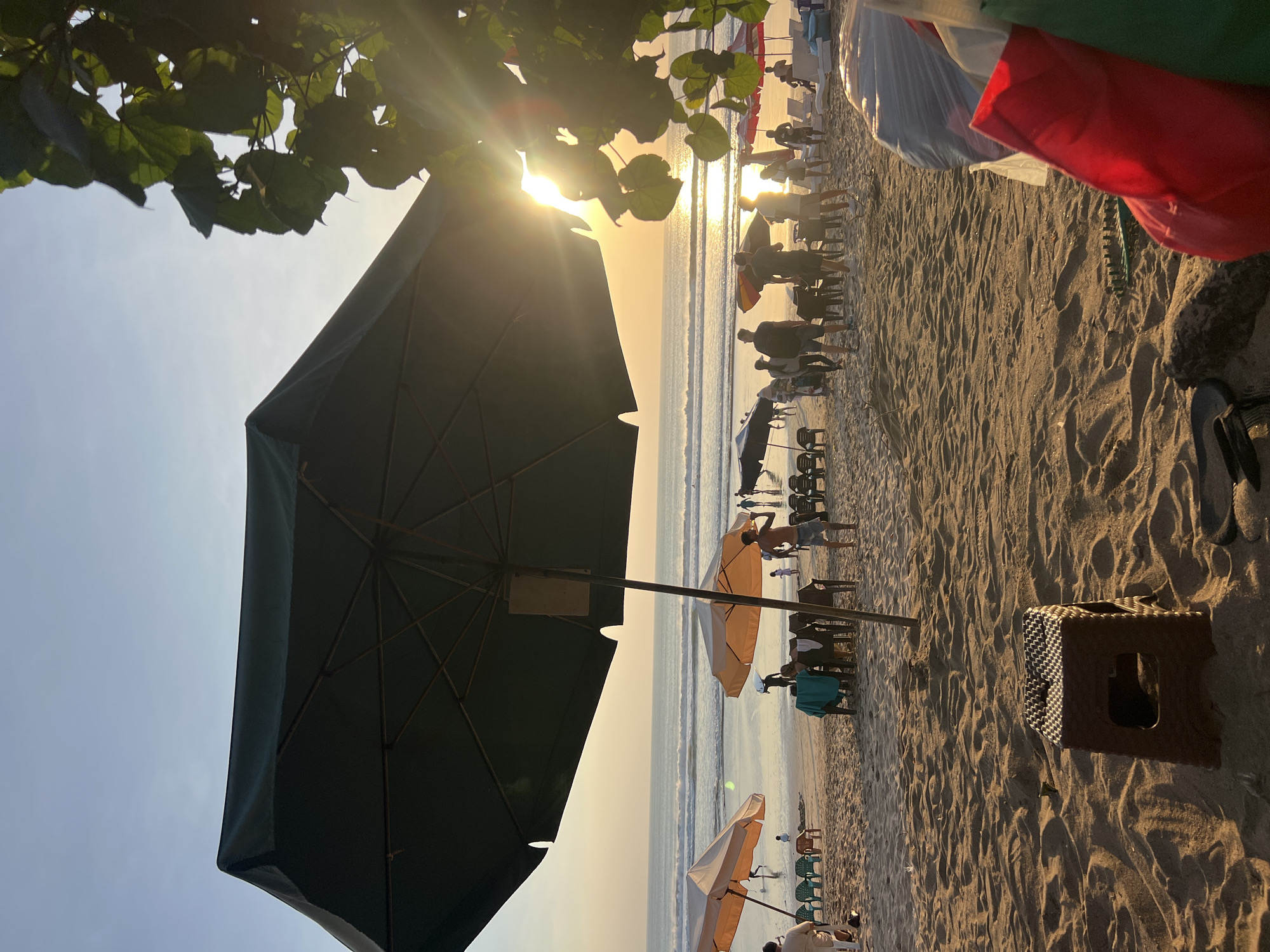 The image depicts a beach scene in Kuta, Indonesia, during sunset. The sun is low on the horizon, casting a warm glow and lens flares across the sky and water. A large, dark green umbrella dominates the foreground, its structure visible beneath the canopy. In the middle ground, several other beach umbrellas, some orange and white, are set up on the sandy beach, with tables and chairs arranged beneath them. People are scattered across the beach, some walking, some sitting, and others gathering. The ocean is calm with gentle waves rolling onto the shore. The sand is textured with footprints and ripples. In the immediate foreground, a small brown plastic stool is visible, along with some colorful bags and flip-flops. Trees with green leaves are visible on the right side of the image, framing the scene and partially obscuring the sun.