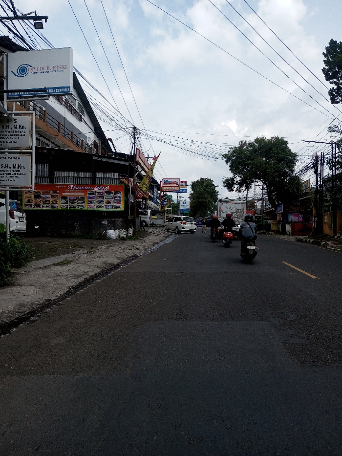 The image depicts an urban street scene under an overcast sky, characteristic of Southeast Asia, contrary to the specified location of Khajuripada, India, as evidenced by visible Indonesian text and brands like "Alfamart" and "Optik B. Riski".
In the foreground, an asphalt road with visible darker patches, possibly recent repairs, extends away from the viewer. A broken yellow lane marking is partially visible on the right side of the road. To the left, an uneven concrete sidewalk with a raised curb is present, bordered by sparse green vegetation. Further left, a multi-story building features a white upper section and a darker ground floor. Attached to this building are several signs: a prominent white sign for "Optik B. Riski" with an eye logo, another sign for "NASKAH KOMPUTER," and smaller white signs displaying text like "S.H., M.Kn." Below these, a large red and white banner displays multiple product images. A white car is partially visible parked near the sidewalk on the far left. Several white sacks are situated at the base of the building.
The midground features three motorcycles traveling away from the viewer on the asphalt road. The closest motorcycle is dark-colored, ridden by a person in a grey jacket and dark helmet. The second is reddish-brown, and the third (furthest) is dark-colored; both are ridden by helmeted individuals. Further down the road, additional vehicles are visible.
On the right side of the street, dense overhead power lines and communication cables are extensively draped across multiple utility poles. A large, leafy green tree stands prominently on the right. Commercial establishments are visible along the right side of the road, including a sign for "Alfamart," a convenience store. The background shows more buildings and trees under the same cloudy sky, indicating a commercial or residential urban environment.