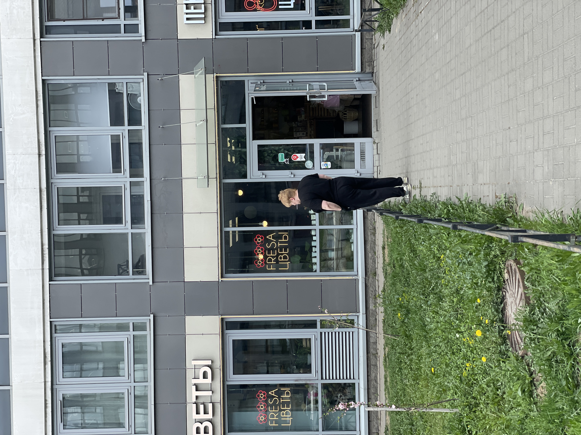 A woman is standing in front of a flower shop. The shop's name, "FRESA ЦВЕТЫ", is visible on the windows. "ЦВЕТЫ" is also written on the side of the building. The building appears to be modern with gray and white panels. A fence runs along the edge of the sidewalk next to a grassy area with dandelions. The person is dressed in black athletic wear. The scene is in an urban setting. Based on the Cyrillic text and architectural style, it is likely Saint Petersburg, Russia.